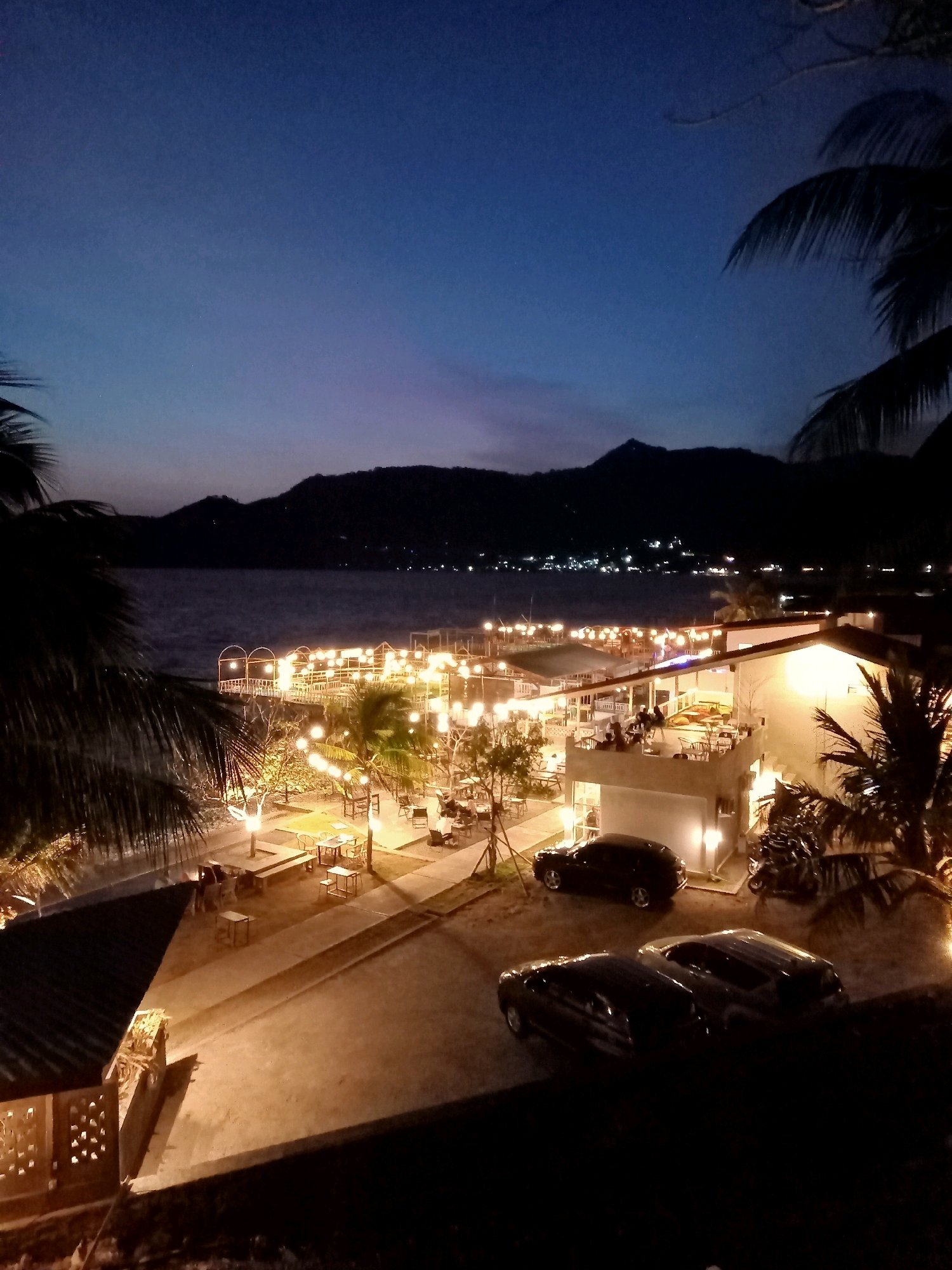 This is a nighttime photograph of a waterfront restaurant or resort in Gorontalo, Indonesia. The scene is illuminated by numerous warm-toned string lights and pathway lights, creating a cozy and inviting ambiance. In the foreground, there are several parked cars, including a dark SUV and two other vehicles. Beyond the parking area, the property features outdoor seating with tables and chairs, some of which are occupied by patrons.
A walkway leads towards a body of water, where a pier or deck area is also lit with string lights. The water is calm and reflects the lights from the shore. In the background, silhouetted hills or mountains rise against a deep blue twilight sky. Palm trees frame the left and right sides of the image, their fronds dark against the fading light. The architecture of the building appears modern, with white walls and visible lighting fixtures. The overall impression is that of a lively establishment enjoying the evening by the water.