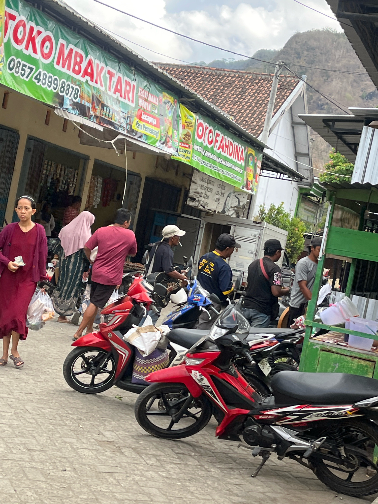 In a street scene in Kendalrejo, Indonesia, a row of storefronts displays banners, including one that reads "TOKO MBAK TARI" with a phone number. People are seen walking and standing near parked motorcycles. A woman in a dark red dress carries a plastic bag and a piece of paper. Another woman in a pink hijab is behind her. A man in a red shirt and black shorts is walking away from the viewer. Several men in casual attire, some wearing caps, are gathered near motorcycles. A green food stall is visible on the right side of the image. The ground is paved with concrete tiles, and a hilly, vegetated landscape can be seen in the background under a cloudy sky.