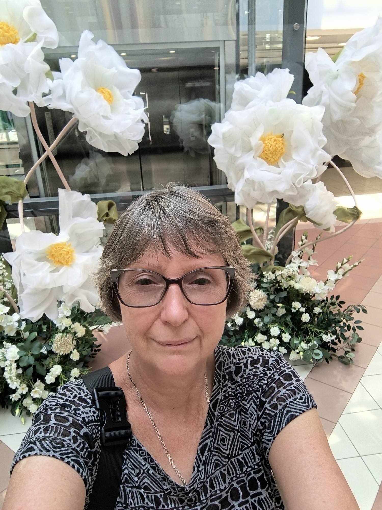 A woman with short gray hair and glasses smiles at the camera in a selfie. She is wearing a black and white patterned shirt and a silver necklace. She has a black strap across her shoulder. Behind her are large, white artificial flowers with yellow centers, and arrangements of smaller white flowers and green foliage. The background appears to be an indoor public space, possibly a mall or atrium, with a glass elevator or entrance visible. The floor is tiled in a light color. The image is taken in Mykolaiv, Ukraine.