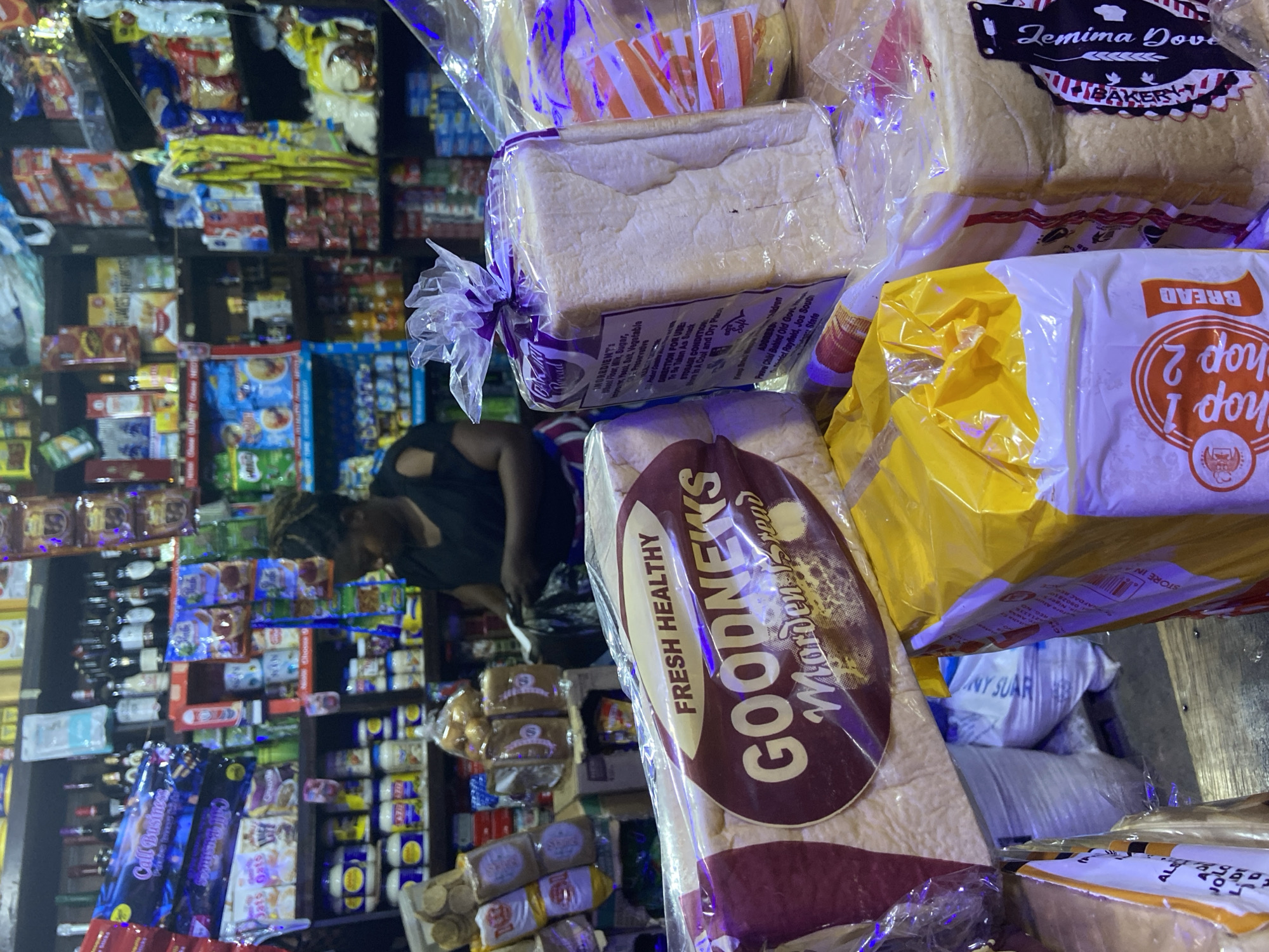 The image depicts a close-up view of a small grocery store or market stall, likely located in Du, Nigeria, given the context. The foreground is dominated by several loaves of bread packaged in transparent plastic. One loaf in the center has a prominent label that reads "GOODNEWS Morden Bread" with "FRESH HEALTHY" above it. To the right, another loaf is wrapped in plastic with a purple ribbon tied at the top, displaying text that includes "INGREDIENTS: Wheat Flour, Milk, Sugar..." and "STORAGE CONDITION: Store in a Cool and Dry Place". To the left and in the background, shelves are packed with various food items, including bottles of beverages, packaged snacks, and jars. A woman with dark hair pulled back is partially visible in the background, appearing to be tending to the store. The lighting suggests an indoor setting, possibly artificial, with a slight blue cast due to lighting effects or image processing. The overall impression is that of a bustling, well-stocked local shop.