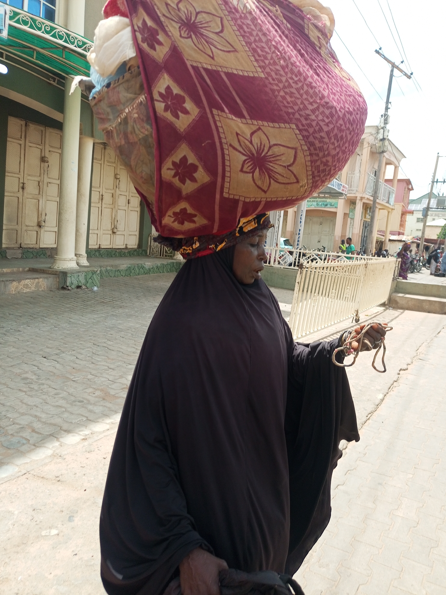 A woman wearing a black abaya and a headscarf balances a large bundle of fabric on her head. The fabric bundle is wrapped in a patterned cloth with floral and geometric designs in shades of maroon and gold. She is walking on a paved walkway in front of buildings. In her right hand, she holds a coiled rope or tether. The background shows a street scene with buildings, a white fence, and a utility pole. The pavement is made of interlocking bricks. The location is identified as Arawa, Nigeria.
