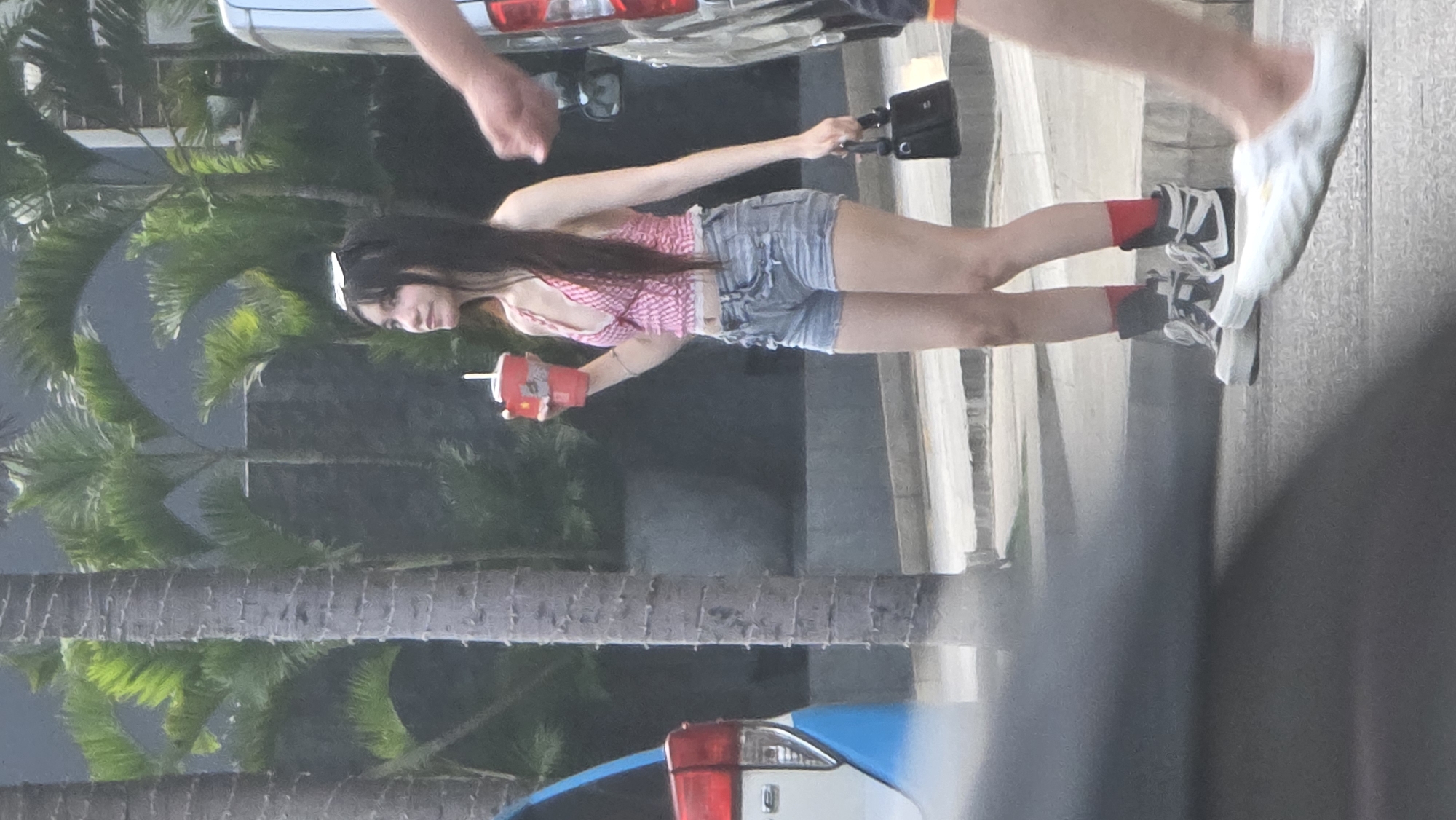 A young woman with long dark hair, wearing a white hairband, stands outdoors in a casual pose. She is dressed in a pink and white checkered crop top, light blue denim shorts, and black and white sneakers paired with red socks. In her right hand, she holds a red McDonald's cup with a white straw, displaying the "McDonald's" logo and "M" symbol. Her left hand holds a small black bag or camera rig. She is looking slightly downwards and to her left.
The background features several lush palm trees with visible trunks and fronds, indicating a tropical setting. Behind the woman, a dark gray building structure is partially visible. On the far left, the rear section of a silver vehicle is seen, and at the bottom, the taillight of a blue car is present. The arm and leg of another person, wearing a white shoe, are partially visible on the right side of the frame. The scene appears to be taking place during the daytime, with soft lighting suggesting an overcast sky or late afternoon. The overall environment conveys a warm, tropical, and urban atmosphere, consistent with Nha Trang, Vietnam.