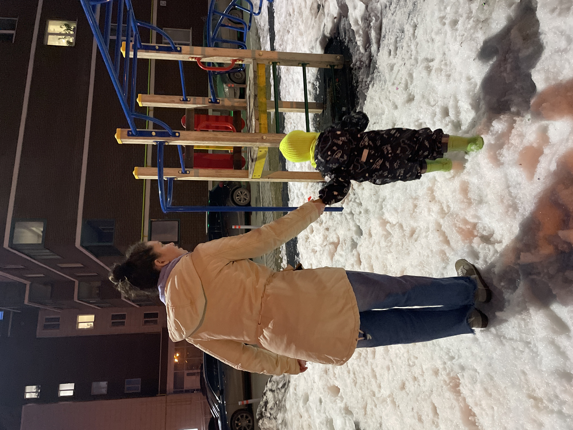 A woman and child are at a playground covered in snow in Izhevsk, Russia. The woman, wearing a tan coat and blue jeans, extends her right arm toward the child. The child is wearing a yellow hat, a patterned snowsuit, and green boots. The playground equipment is made of blue and wooden bars and ladders. A multi-story building is visible in the background. Cars are parked along the side of the building and near the playground.