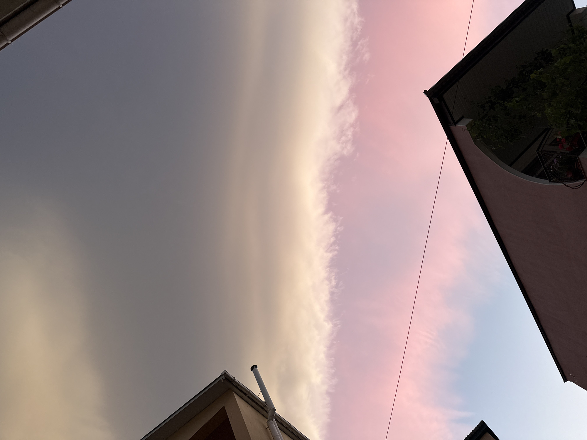 The image shows a sky with a distinct division between gray, overcast clouds and pinkish-blue, clear sky. The photo is taken from a low angle, looking up between buildings. Building structures are visible in all four corners of the frame. A house is in the lower left and a building in the upper right. Location: Unknown, Unknown.