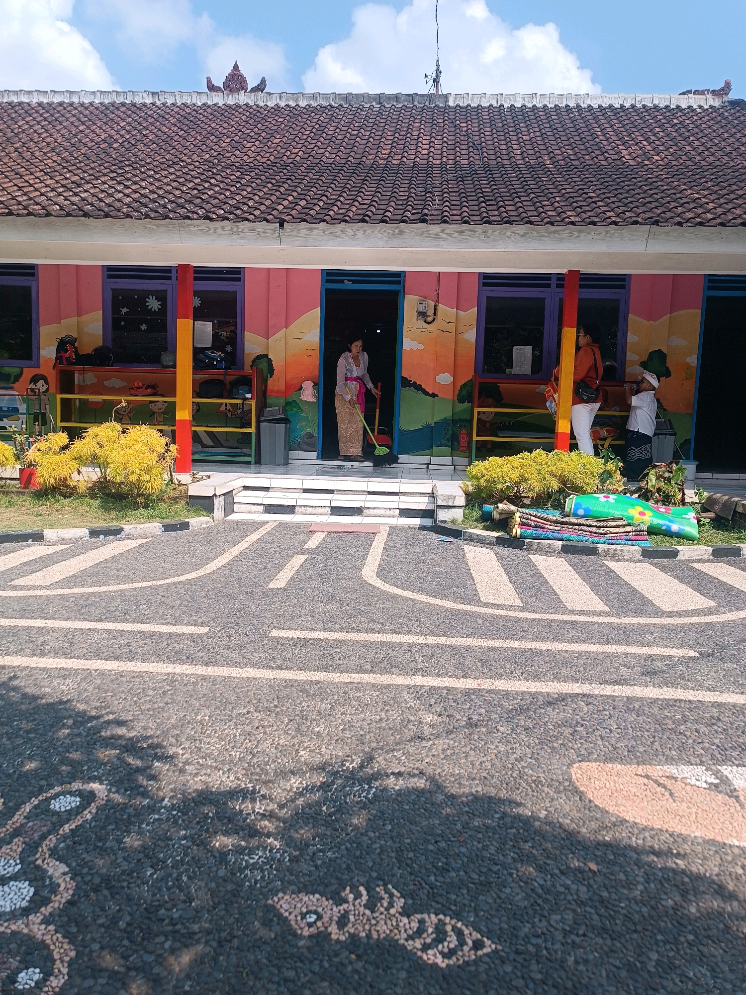 A woman in a traditional sarong and white top sweeps the entrance of a colorful building with a broom. The building appears to be a school or community center, with brightly painted murals of mountains and children on its facade. A yellow, red, and blue striped pole supports an awning over the entrance. Shelves with school supplies are visible inside. To the right of the entrance, a woman in an orange jacket and white pants stands near a child in a white shirt and a cap. Several rolled-up mats are stacked on the ground near the entrance. The ground in front of the building is paved with dark stones and white lines marking a roadway, suggesting an outdoor play area or a road safety education space. The area is surrounded by greenery, and the sky is blue with scattered clouds. The building has a tiled roof with decorative elements at the peak. The scene is set in Tabanan, Indonesia.