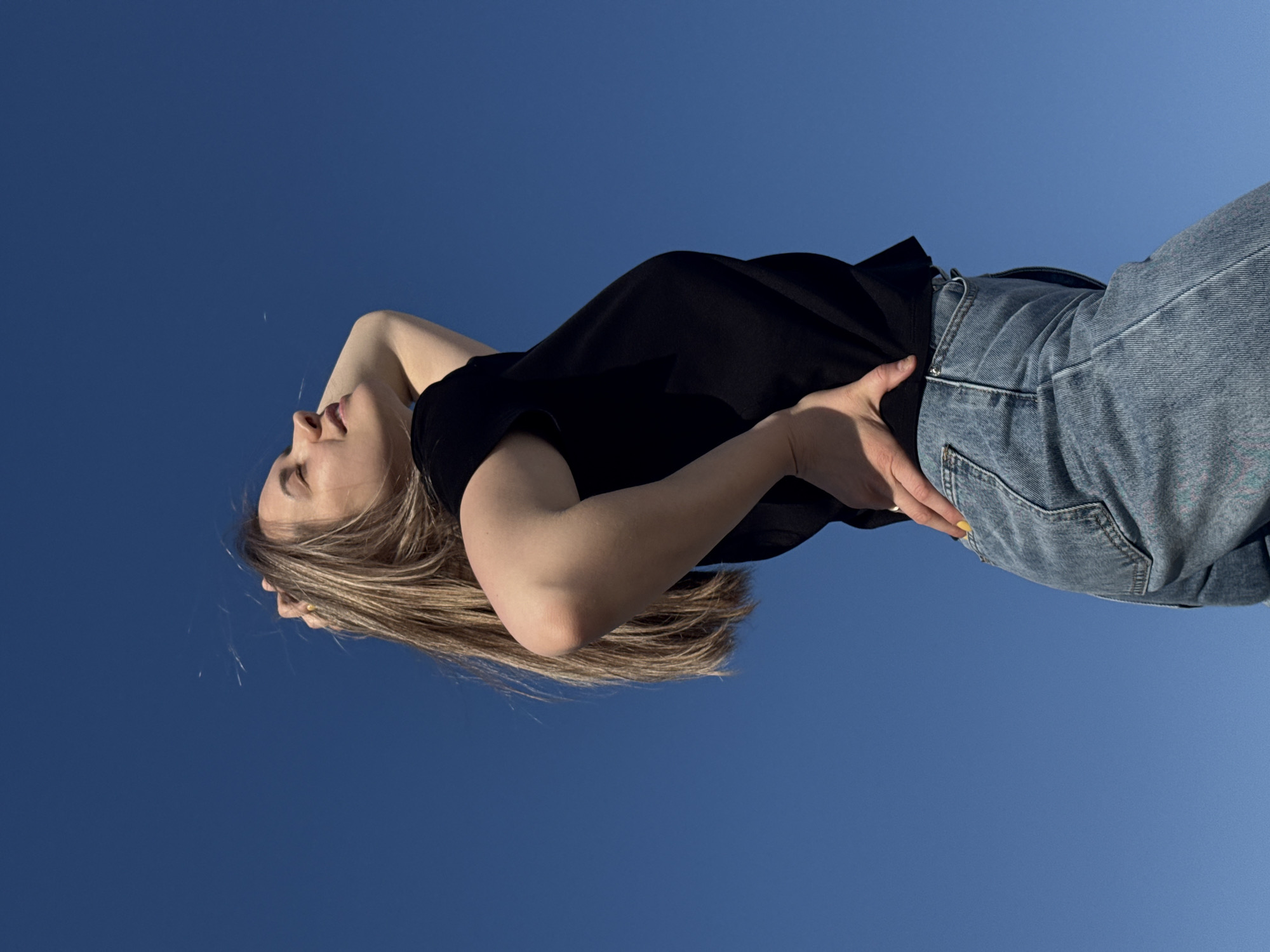 A young woman with blonde hair is seen from a low angle, against a bright blue sky. She is wearing a black top and blue jeans. Her head is tilted back, with her hands positioned near her head and waist. The exact location within Chaikovsky, Russia, is not discernible from the image.