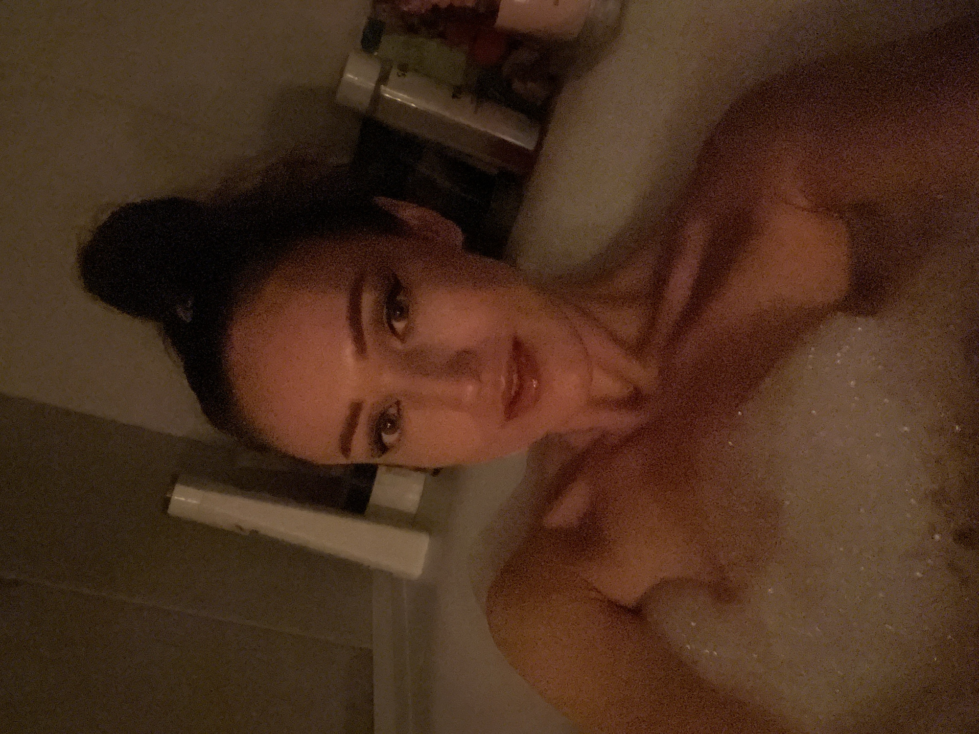 The image shows a woman in a bubble bath. She is lying in the bathtub, with her head resting on the edge. She has her hair tied up in a bun. She is wearing makeup, including eyeliner and lipstick. In the background, there are several bottles of toiletries on the edge of the bathtub. The scene appears to be in a bathroom. There is no information in the image to suggest the location is in the city of Kazan, Russia.