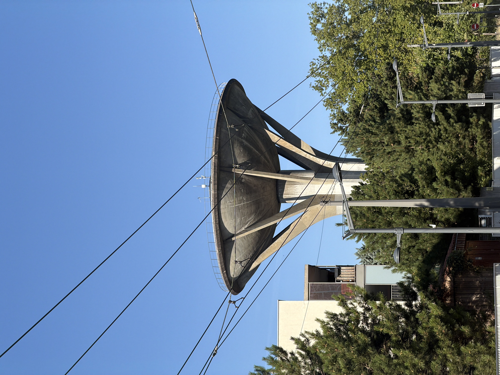 The image shows the iconic water tower of Fort de Vaise, located in the 9th arrondissement of Lyon, France. The concrete structure, with its distinctive mushroom-like cap supported by angled pillars, dominates the upper portion of the frame. Several power lines crisscross the sky in front of the water tower, creating a striking geometric pattern against the clear blue sky. To the left of the water tower, a modern building with a beige facade and balconies is partially visible, surrounded by lush green pine trees. In the foreground, more trees, streetlights, and a small wooden shed add to the urban landscape. The scene appears to be captured on a sunny day, with clear visibility and sharp shadows.