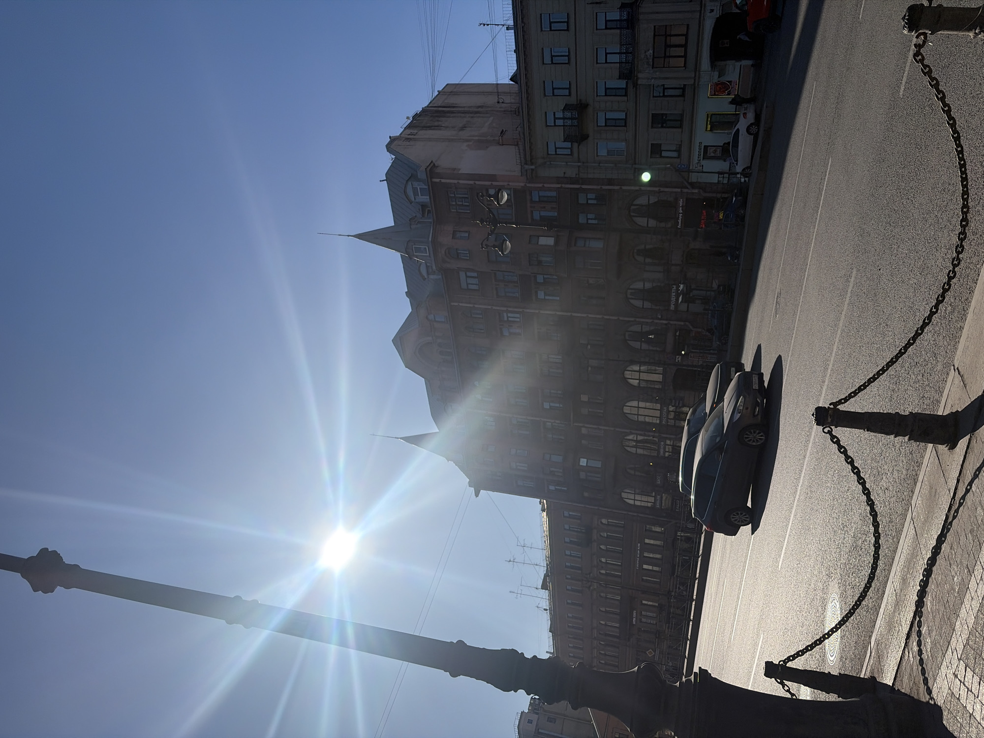 The image shows a street scene in Saint Petersburg, Russia, likely taken on a sunny day. The camera is oriented upwards, capturing the sky, the top portion of a building, part of the road, and foreground elements like a lamp post and chain railing.
Building: An ornate, multi-story building with several rows of windows is visible. The architectural style appears to be pre-Revolutionary. The building's color is a muted tan/brown.
Street: The road is paved, and at least two dark-colored cars are parked.
Sky: The sky is bright blue with intense sun flares radiating from the position of the sun.
Foreground: A cast-iron lamp post and a chain railing stand in the foreground, partially obscuring the view. The lamp post and chain fence create shadows on the sidewalk.