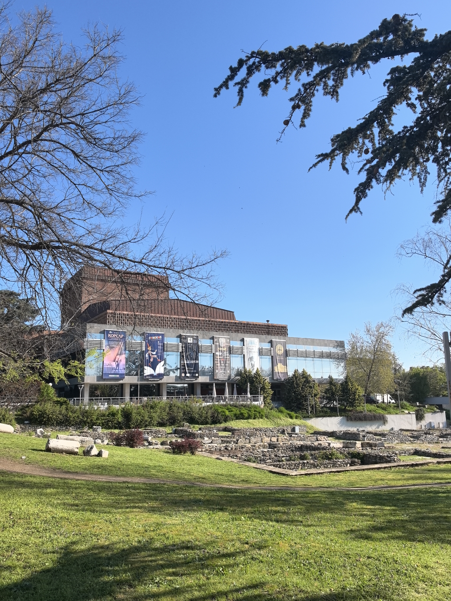 The image depicts a modern building with large banners hanging on its facade, likely a theater or cultural center, in Stara Zagora, Bulgaria. In front of the building is a partially excavated Roman ruins site with stone foundations visible among green grass. Trees surround the scene, with bare branches and full foliage. The sky is clear and blue.