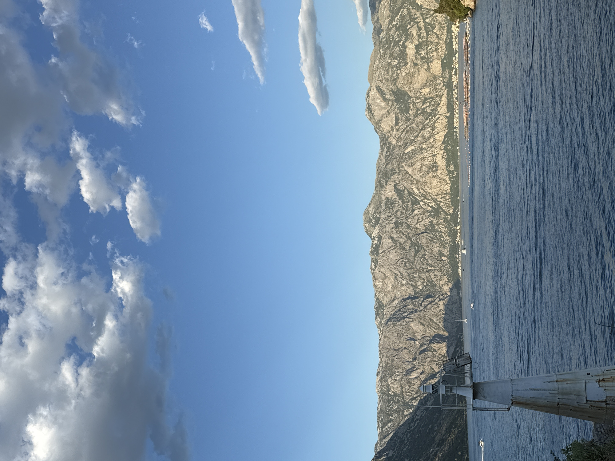 The image shows a wide view of a bay with a mountainous backdrop, likely in the Kotor Bay area of Montenegro, possibly near Đurići. In the foreground, to the left, stands a white, weathered lighthouse. The bay is a deep blue, with ripples on its surface. Several boats are visible on the water, including a white speedboat to the far left, and sailboats further out. In the middle distance, there are some structures on the water that appear to be floating platforms or aquaculture farms, with a town or village situated along the shoreline at the base of the mountains. The mountains themselves are rugged and rocky, with sparse vegetation. The sky is a bright blue with scattered white and grey clouds, suggesting it is daytime. The lighting indicates it might be late afternoon or early morning, with shadows cast by the mountains.