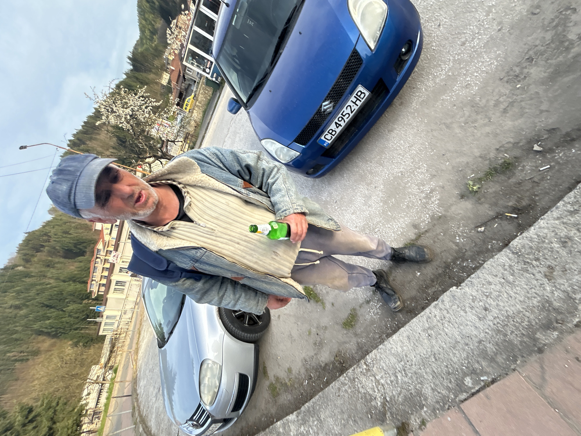 A man in a denim jacket and cap holds a green bottle near a blue Suzuki Swift car with license plate "CB 4952 HB." He stands near a silver car and a curb, with buildings and trees visible in the background. The location is Zlatograd, Bulgaria.
