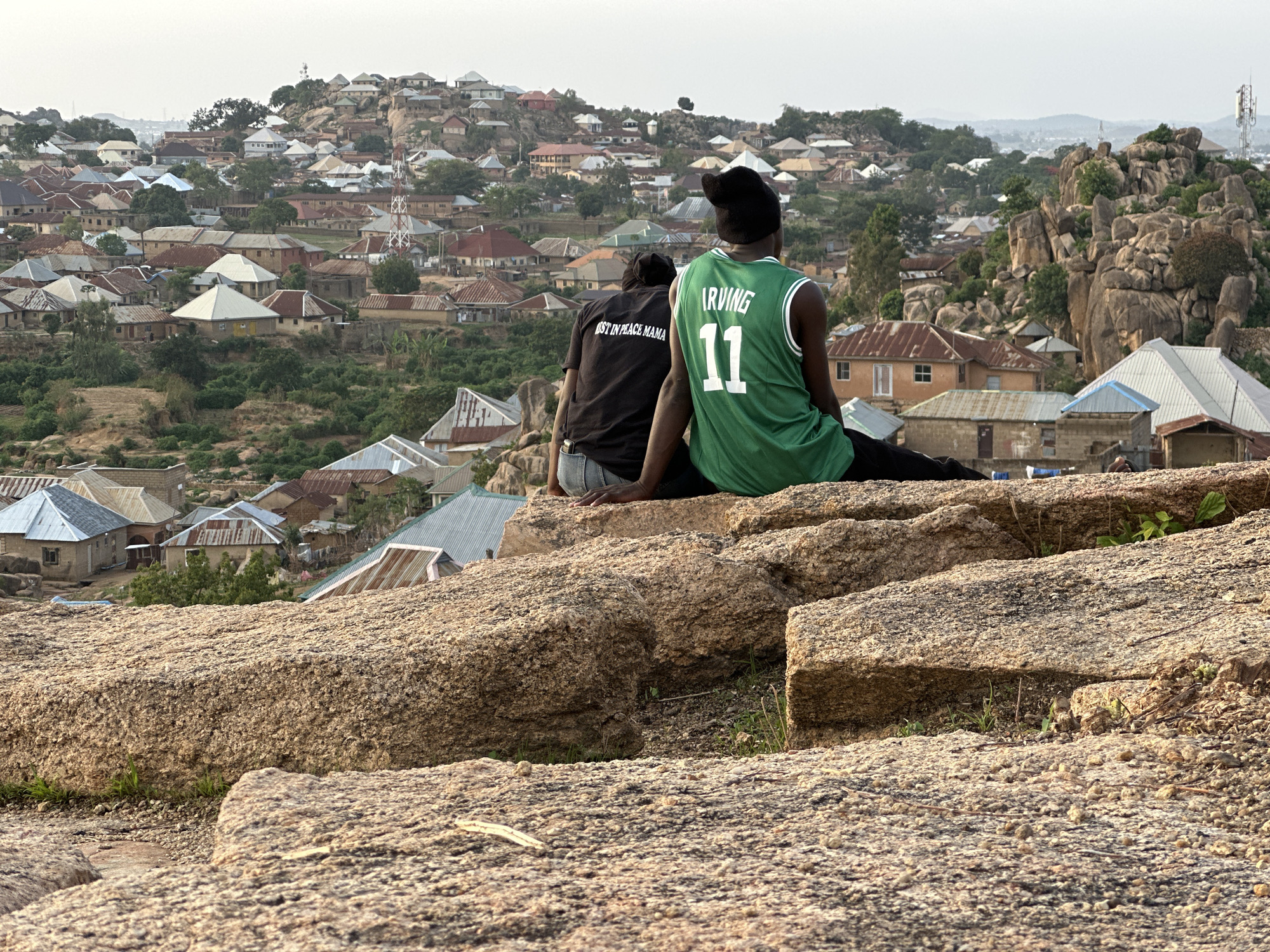 Two individuals sit on a rocky outcrop overlooking a dense residential area in Jos, Nigeria. The person on the right wears a green basketball jersey with the name "IRVING" and the number "11" on the back, with a black beanie. The person on the left wears a dark t-shirt with the words "REST IN PEACE MAMA" printed on the back.
The houses below have predominantly corrugated iron roofs. The landscape includes a mix of residential buildings and scattered trees. In the background, there are rock formations, and on top of the highest point is a group of buildings with antennas. The overall scene is bathed in warm light.