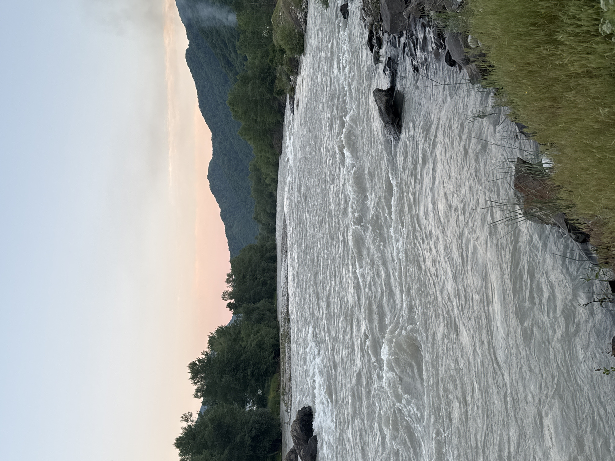 This is a landscape photograph taken in Kamenomostsky, Russia. The image depicts a wide, fast-flowing river with turbulent, white water. The riverbanks are lined with dark, wet rocks and lush green grass. In the background, a dense forest covers the slopes of a large mountain range. The sky above is a pale, hazy gradient, transitioning from a soft pinkish hue near the horizon to a light blue at the top, suggesting either sunrise or sunset. Some light mist or clouds are visible near the mountain peaks. The overall scene conveys a sense of wild, natural beauty and tranquility, characteristic of a mountainous, riverside setting. There is no visible text in the image.