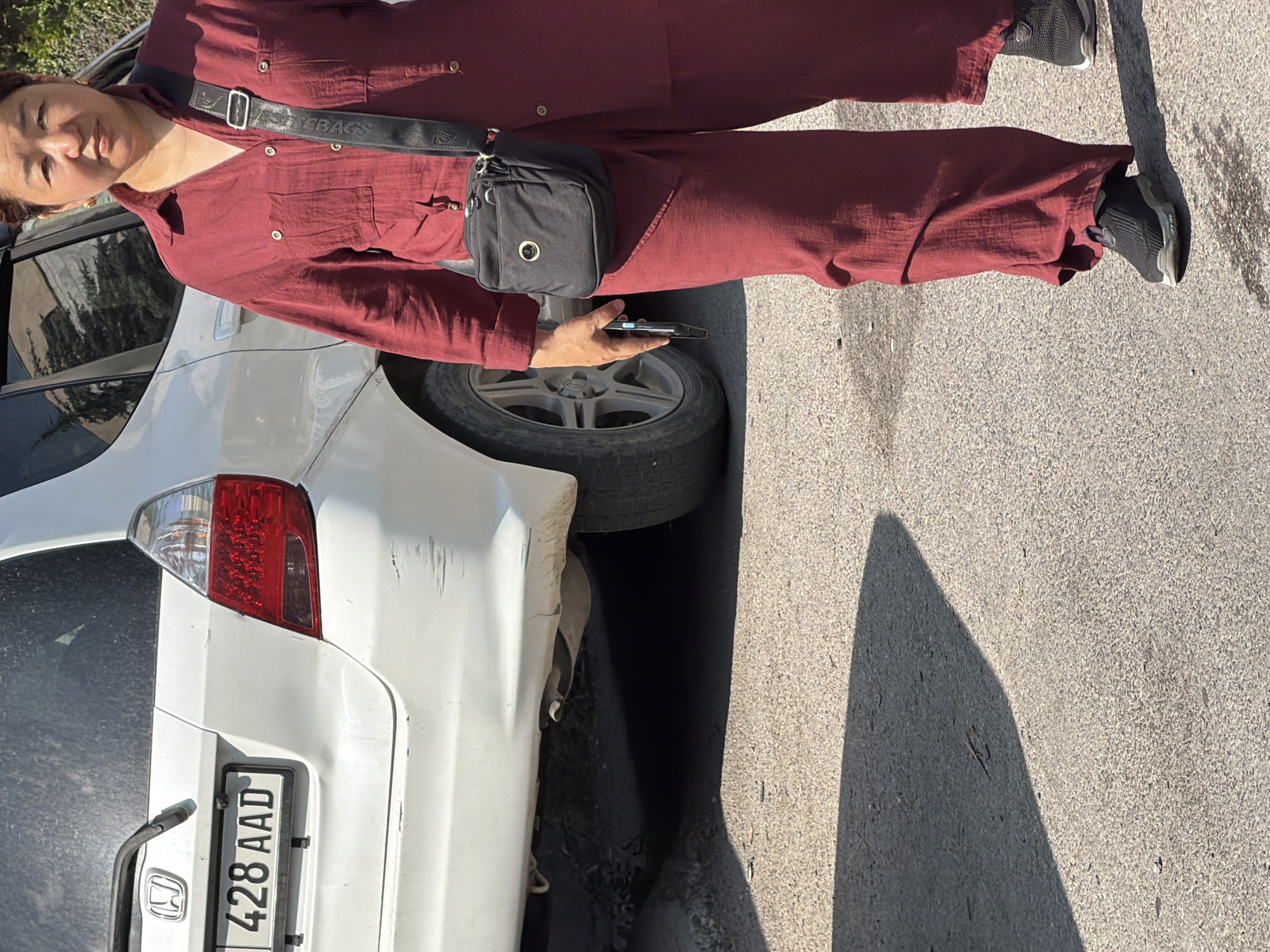 A medium shot captures a woman from the knees up, standing next to a white car. She is wearing a loose-fitting, maroon-colored jumpsuit and black sneakers. A small black crossbody bag is slung over her right shoulder, and she holds a smartphone in her left hand. The car, a white hatchback, is parked on an asphalt surface, its rear bumper slightly dented, and its license plate reads "428 AAD." The background shows a dimly lit area beneath the car. The scene is outdoors, under bright daylight, with a long shadow cast on the ground.