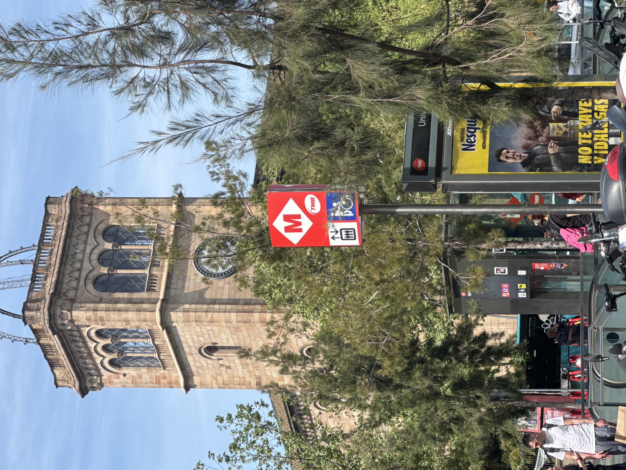 A daytime street scene in Barcelona, Spain, features a mix of historical architecture, urban infrastructure, and natural elements under a clear blue sky. On the left, a tall, ornate stone tower, possibly part of a university or church, dominates the skyline with its arched windows and visible clock face. Lush green trees with needle-like leaves are scattered throughout the frame, partially obscuring the building and other street elements.
In the mid-ground, a prominent red and white "M" symbol Metro sign on a pole clearly indicates a subway entrance. This sign also displays "TMB" (Transports Metropolitans de Barcelona), accessibility symbols (wheelchair, escalator, stairs), and metro line indicators "L1 L2". Another sign on the same pole reads "TMB" and "Univ," likely referring to the Universitat metro station.
To the right, a large advertising billboard for "Nestlé Nesquik" is visible, showing a smiling man pointing at the product. Text on the billboard includes "Nestlé Nesquik HERMANOS" and the Spanish phrase "NO TE RAYES, VIDALEGAS." The frame of the billboard also shows "JCDecaux."
Several people are present, including a man in a white t-shirt on the far right, standing near parked motorcycles or scooters. Other figures are partially visible in the background, suggesting a busy urban environment. The sunny weather and bright natural light contribute to the lively atmosphere. The scene captures daily life around a metro station and a significant architectural landmark.