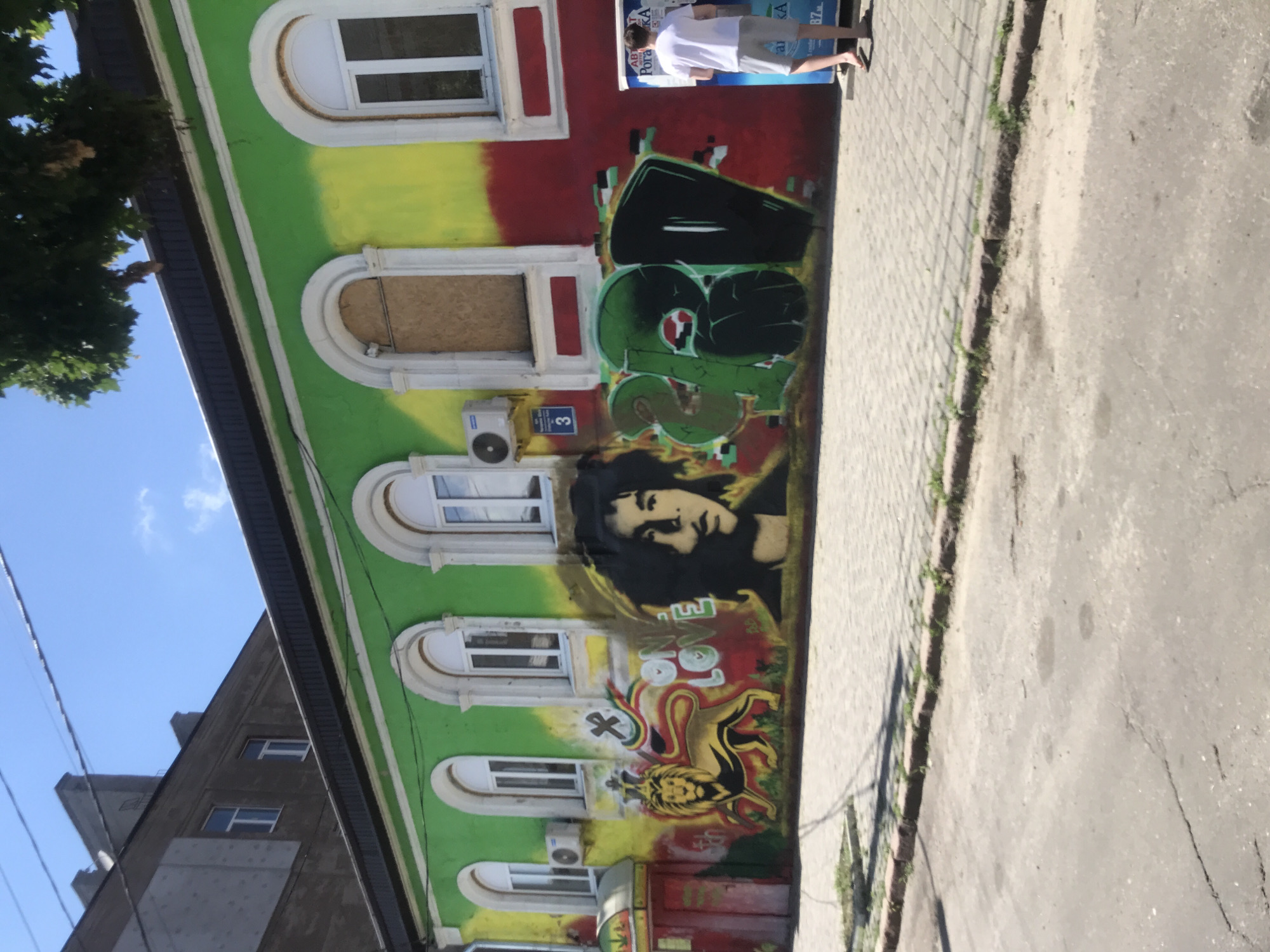 The image shows a building facade adorned with vibrant graffiti, featuring bold red, yellow, and green colors. On the left, a stylized lion with a crown and reggae-themed colors is depicted. To its right, the words "ONE LOVE" are painted in bubble letters. A black and white portrait of a person with prominent dreadlocks occupies the central portion of the mural. To the right of the portrait, a large, graffiti-style tag in green and black dominates the wall.
A person wearing a white t-shirt and grey shorts stands on the right side of the image, facing away from the camera and looking at an information kiosk. The kiosk has Cyrillic lettering and a digital display. The building has arched windows, some of which are boarded up. An air conditioning unit is mounted on the wall to the left of the windows. The foreground shows a paved sidewalk and a cracked asphalt street. Overhead, power lines are visible against a clear blue sky. The environment suggests an urban setting.