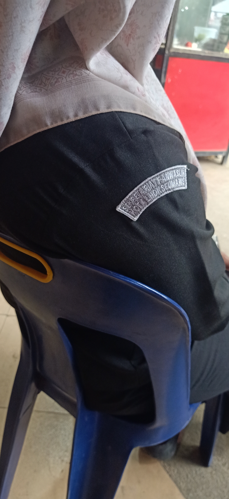 The image shows a close-up view of a person's back, seated on a blue chair. The person is wearing black pants and a light-colored headscarf with a subtle floral pattern. A white embroidered patch is visible on the left side of the black pants. The patch has curved text that appears to read "SEKRETARIAT WANWASLIH KOTA LHOKSEUMAWE".
The setting appears to be indoors, possibly a cafe or a public waiting area, given the presence of a red structure in the background which could be part of a counter or display. The flooring is tiled. The lighting suggests it is daytime. The focus is on the clothing and the patch, hinting at an organizational affiliation. The image is taken from a slightly elevated angle, looking down at the person's back.