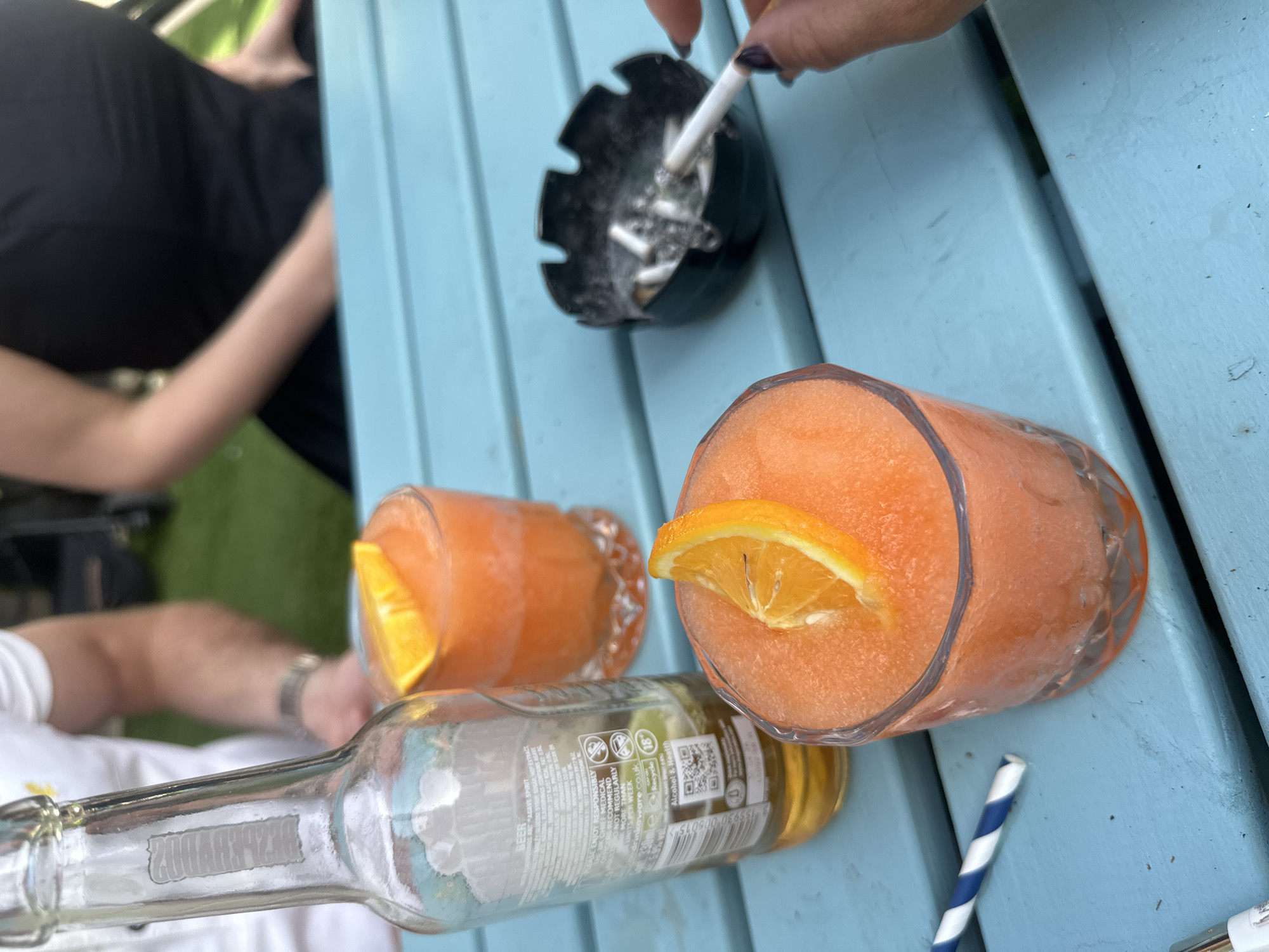 The image depicts a light blue outdoor table scene. On the table are two glasses filled with an orange slush-like beverage, each garnished with a slice of orange. An empty glass beer bottle is laying on its side next to the glasses. A blue and white striped straw sits near the drinks. In the upper-right corner, a hand holds a lit cigarette over a black ashtray filled with cigarette butts. In the background, parts of people are visible, including a person in a wheelchair, suggesting an outdoor patio or garden setting. The OCR mentions UK, specifically that UK's chief medical advises of responsible drinking. This does not pinpoint this scene to London.
