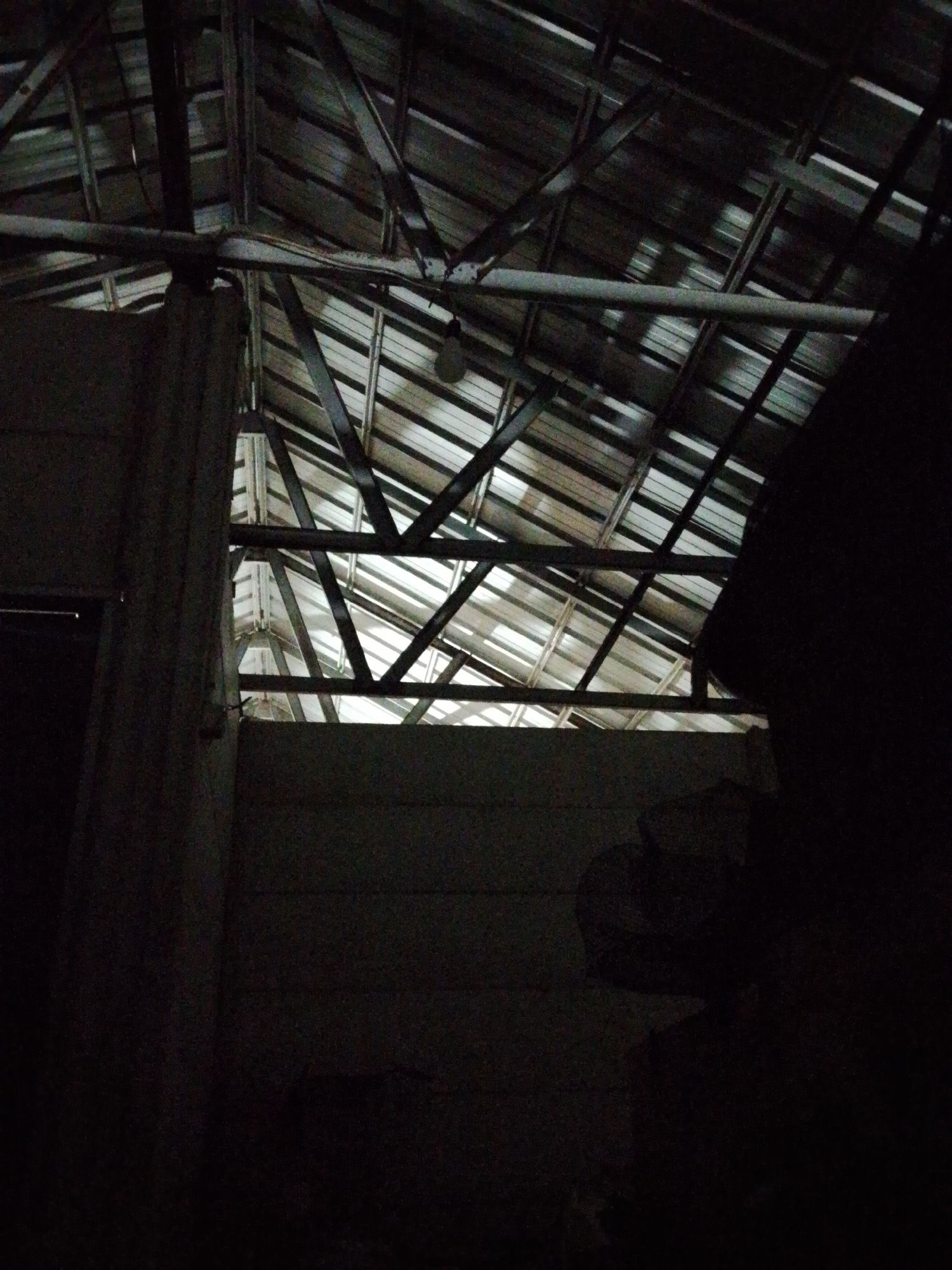 The image depicts the interior of a building in Bukit Ketok, Indonesia, possibly a warehouse or storage space, illuminated by natural light filtering through the roof. The roof has a complex structure of metal beams and corrugated metal sheets. The lower portion of the building is very dark, obscuring details of the walls and objects within. A partial wall is visible on the left side.