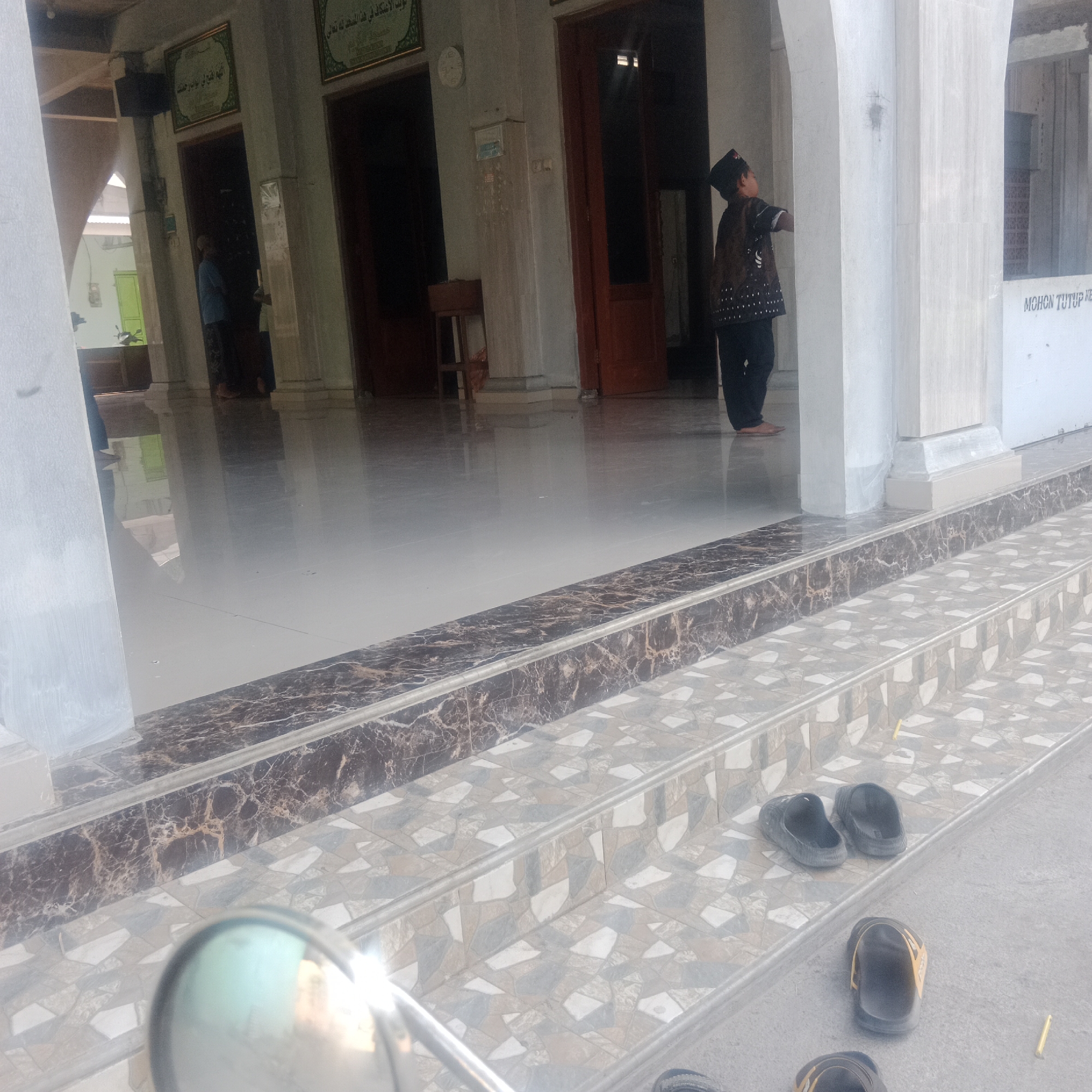 The image depicts the exterior of a building with a tiled staircase and a polished, reflective floor. A young boy wearing a dark cap and a patterned tunic is visible in the background, facing away from the camera and looking upwards. To the left, another person in a blue shirt is partially visible near a doorway. Several pairs of black and orange sandals are placed on the steps and the ground. The environment suggests a public space, potentially a place of worship or community gathering, given the architectural style and the presence of religious text in an overhead frame. The lighting indicates it is daytime. The location is identified as Tegowanu Kulon, Indonesia.