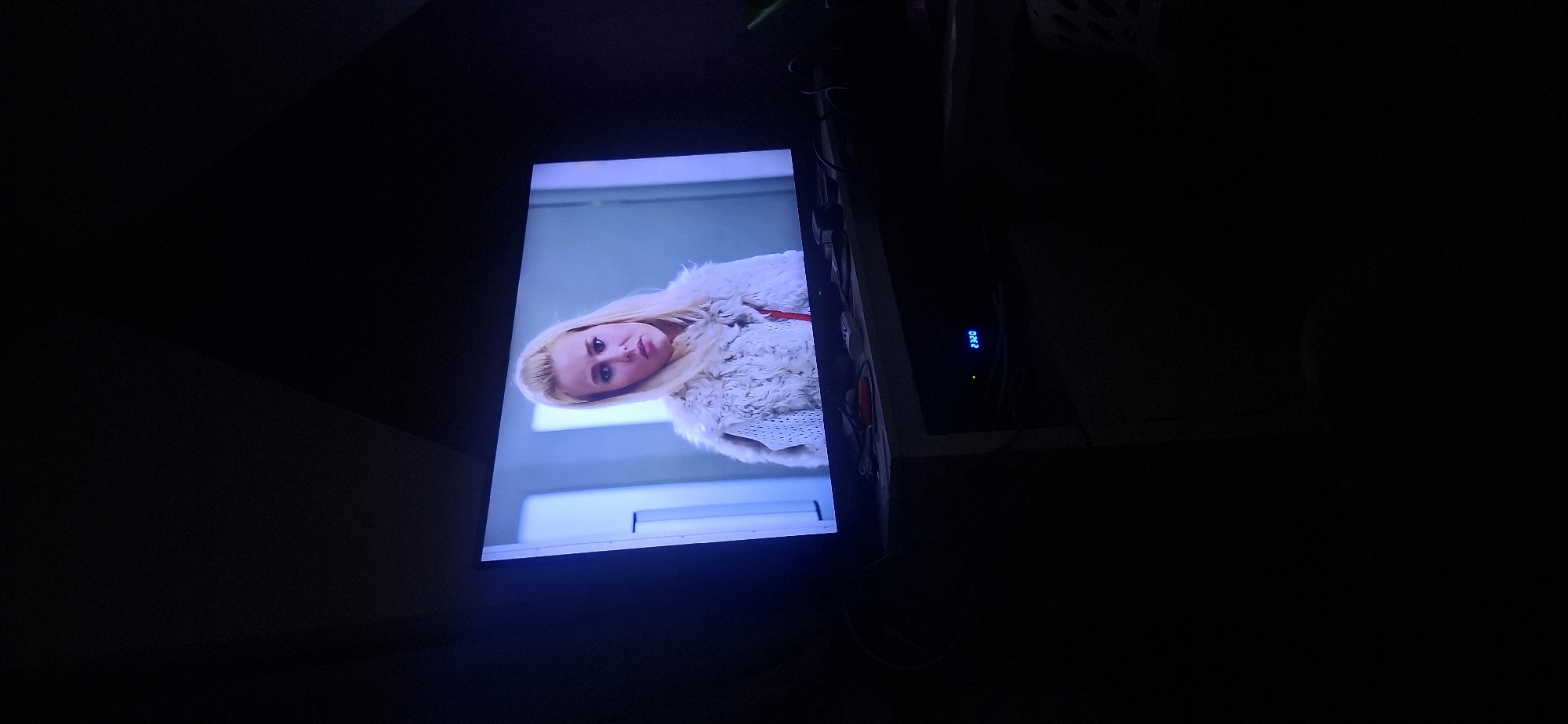 A woman with blonde hair and a white furry coat is standing in front of a blue-tinted wall. She is wearing a red accessory around her neck. The image is taken from a low angle, showing a television screen displaying the woman. Below the television, a black entertainment center is visible with a blue digital display showing "23:20". The overall scene is dark with the television screen being the primary source of light. The location is identified as Yeroham, Israel, however, there are no specific landmarks to confirm this.