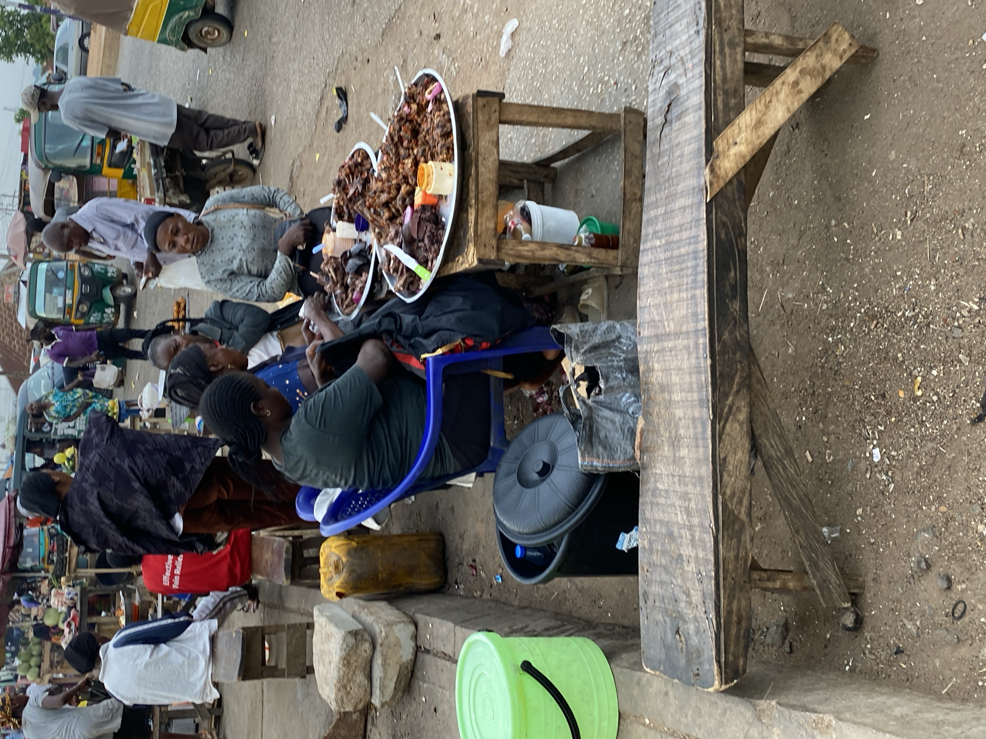 Bukuru, Nigeria. Street scene featuring a gathering around a food vendor. Two large platters filled with grilled meat sit on a small wooden table. A woman in a green shirt sits on a blue plastic chair, facing the food. Other individuals are seated or standing nearby. A wooden table with a makeshift leg is visible in the foreground. A green bucket sits near the curb. Vehicles, including a tricycle, are in the background.