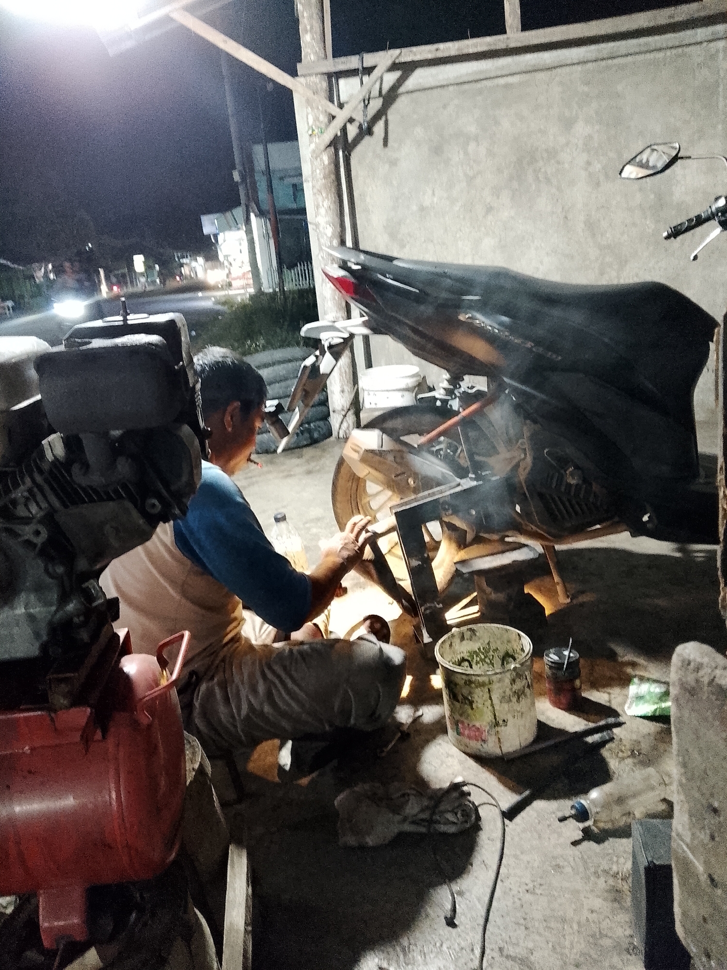 A man is seen working on a motorcycle at night in what appears to be an open-air workshop or garage in Geulanggang Teungoh, Indonesia. The scene is illuminated by a bright overhead light. The man, wearing a blue and white shirt and light-colored pants, is crouched down and appears to be grinding or cutting a metal part of the motorcycle. Sparks are visible, indicating metalwork is in progress. A black motorcycle is partially visible, with its rear section prominently featured. In the foreground, a red air compressor and a large, weathered bucket containing green plant matter are present. Various tools and other objects are scattered around the workspace, including a plastic bottle and a power cord. The background shows a concrete wall and a glimpse of a street scene with car headlights, suggesting the workshop is near a road. The overall impression is one of active repair work in a functional, albeit somewhat cluttered, environment.