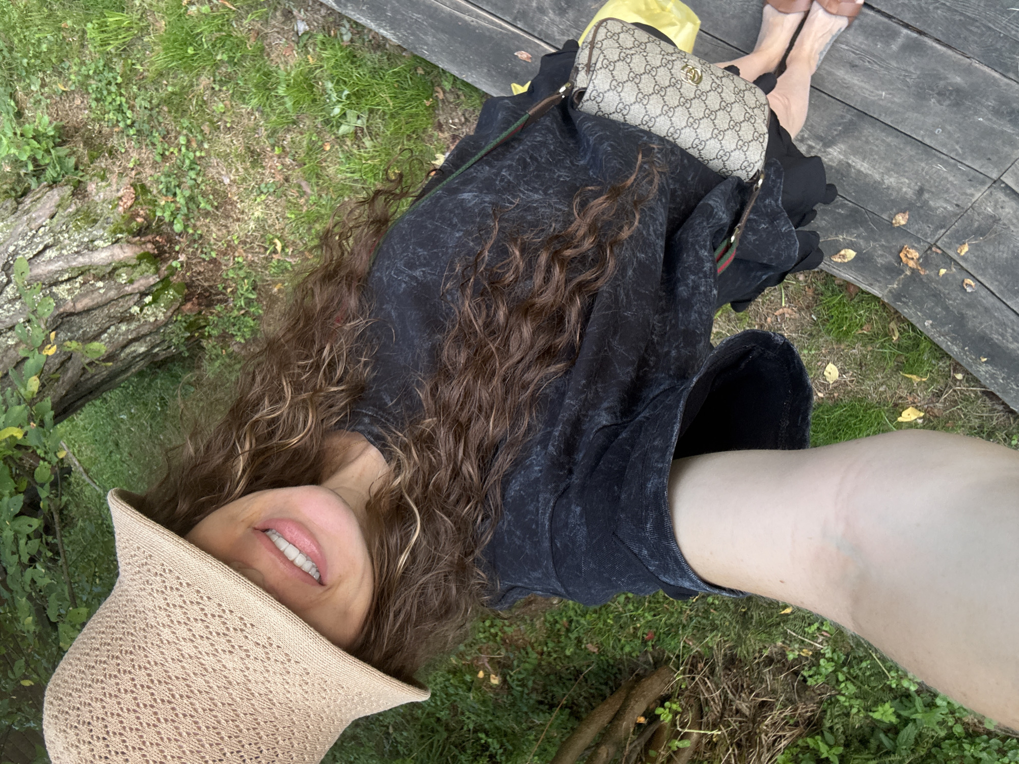 The image shows a woman lying down outdoors, with her face and upper body visible. She is wearing a straw hat that partially covers her face, and she is smiling. Her hair is long and curly, and she is wearing a dark-colored top. A Gucci-branded purse is visible near her, along with a yellow bag. Her legs are also visible, and she is wearing open-toed shoes. The background consists of grass, trees, and a wooden surface, suggesting an outdoor setting such as a park or a garden. The lighting indicates it is daytime, and the overall mood seems relaxed and casual. There is no visible text in the image.