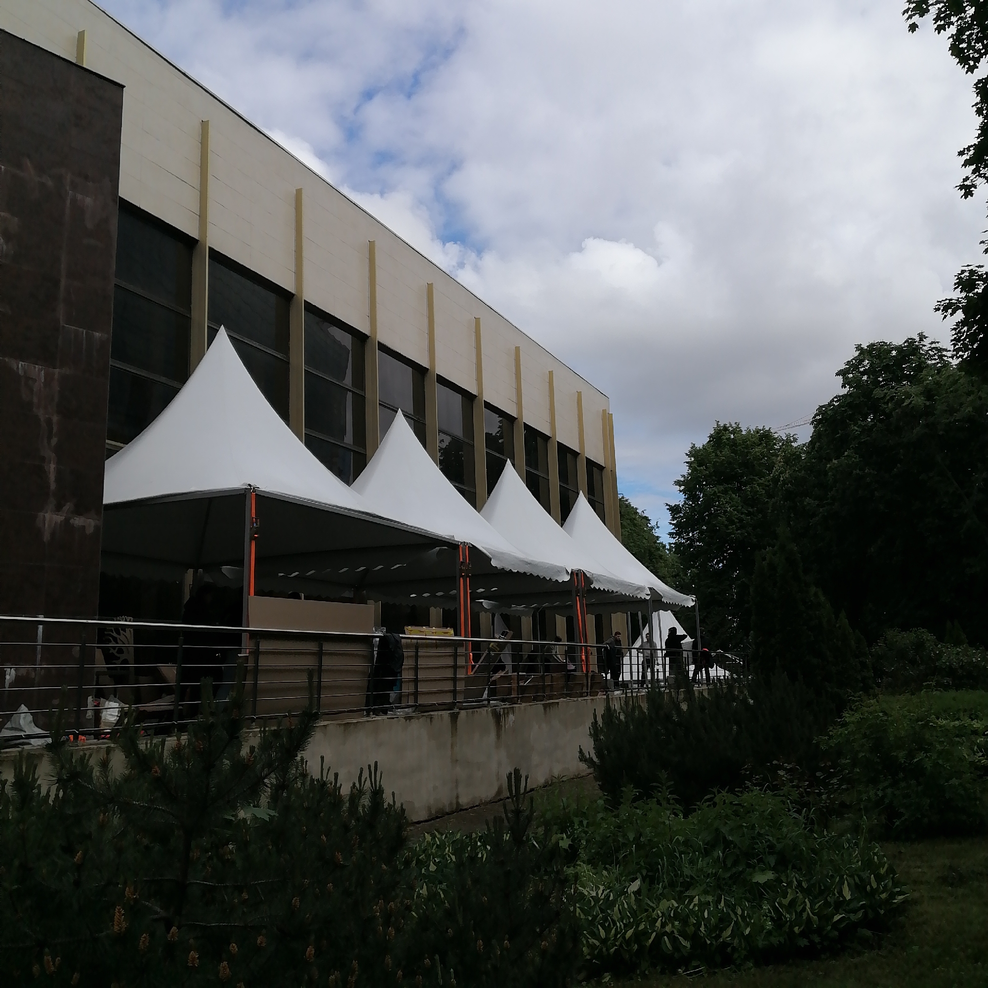 The image shows the front of a building in Moscow, Russia. The building has a light-colored facade with large windows and vertical beige columns. Four white tents with pointed tops are set up in front of the building, along a raised patio area enclosed by a dark metal railing. People can be seen under the tents, possibly setting up for an event. The foreground features landscaping with various plants and bushes. Behind the building and to the right, there are trees and a cloudy sky.