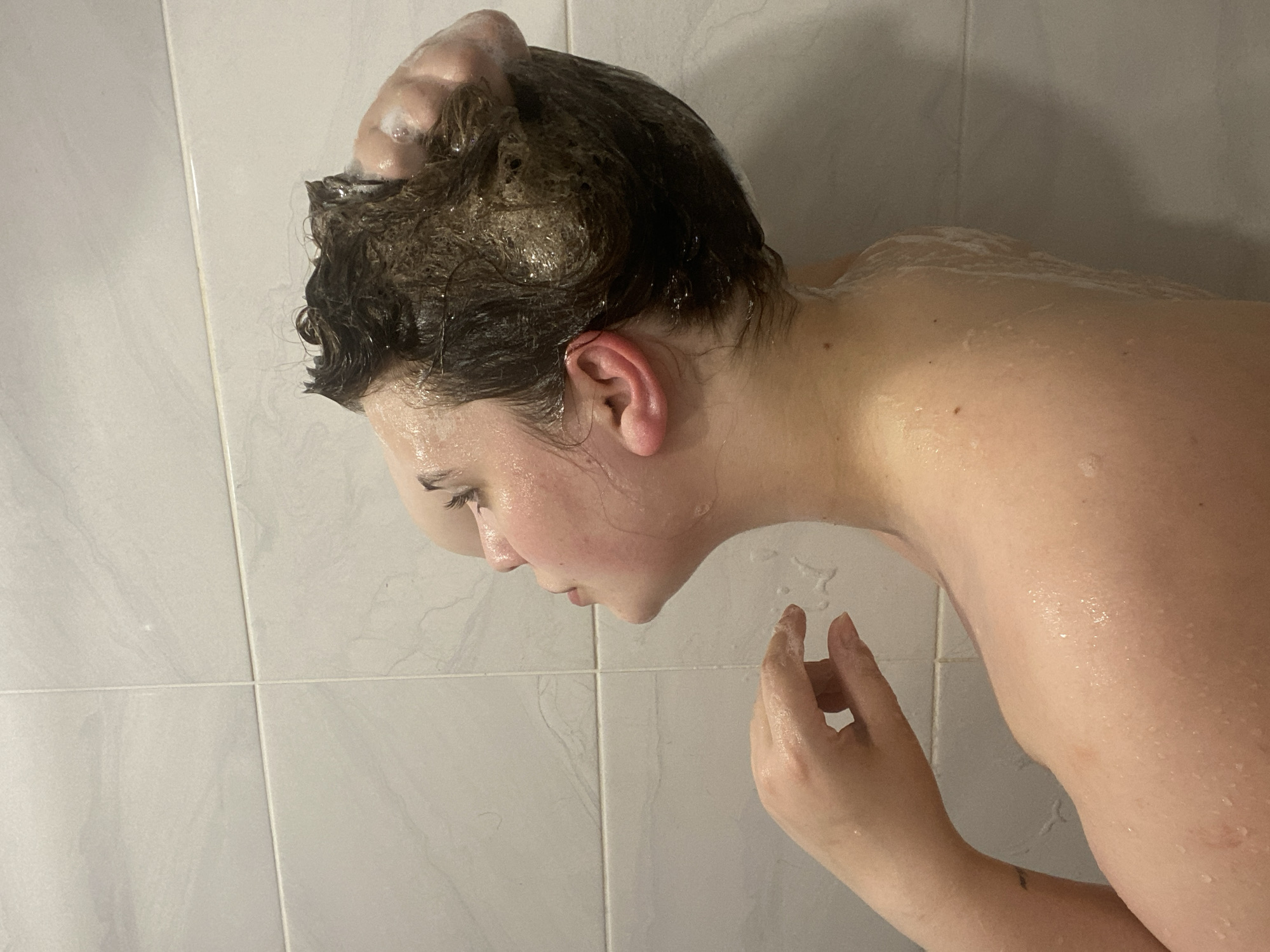 The image captures a close-up, side view of a person engaged in personal hygiene, specifically washing their hair and body. The individual, seen from the upper back and the right side of their face, has dark brown hair thoroughly covered in white shampoo or soap foam. Their skin is wet, with suds also visible on their shoulders and upper back. The person is leaning forward, with their head tilted downwards, a common posture for washing hair in a shower. A hand is visible near their chin and neck, also wet. The background is composed of white, rectangular tiles, suggesting an indoor setting like a bathroom or shower stall. The overall scene conveys a private moment of routine self-care. No specific time of day or emotional expression is evident, and there is no visible text. The provided location context is Chernushka, Russia, though no specific visual cues within the image confirm this.