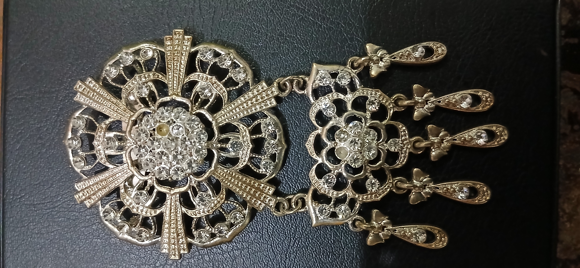 The image shows a close-up, top-down view of a vintage-style brooch. The brooch is ornate, featuring a floral design with numerous clear rhinestones embedded in a gold-toned metal setting. The top portion of the brooch is a large, circular, flower-like shape with radiating petal details and a central cluster of rhinestones. Dangling from this top section is a smaller, similar floral motif, also adorned with rhinestones. Attached to the bottom of this second floral piece are five teardrop-shaped pendants, each with a small rhinestone at its center and delicate filigree detailing. The brooch is placed on a dark, textured surface, likely leather, which appears to be a dark blue or black. The lighting is somewhat uneven, creating subtle shadows around the brooch. No specific location within Samarinda, Indonesia is identifiable from the image itself.