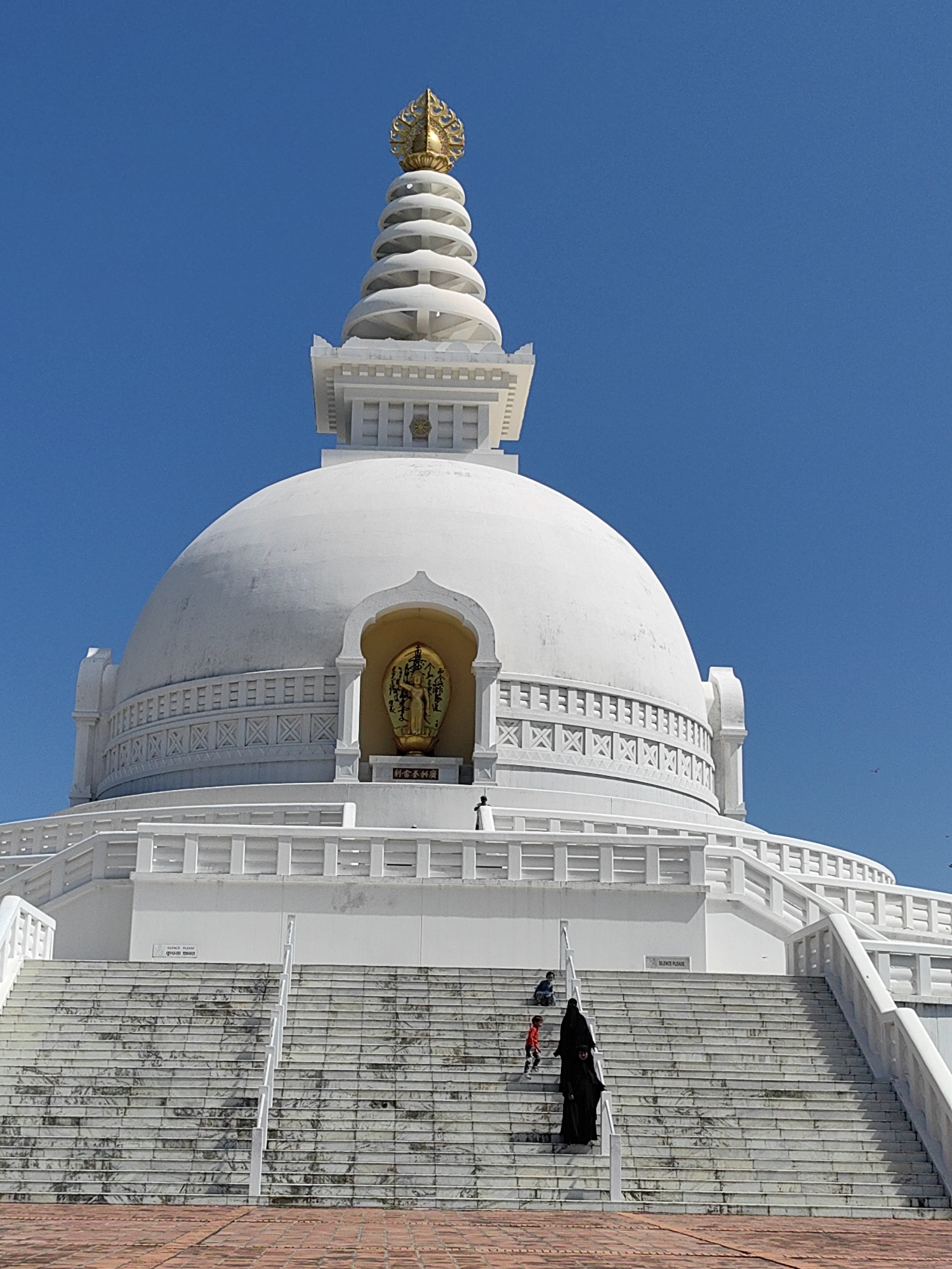 In the city of Parsa, Nepal, a white stupa with a golden finial stands against a clear blue sky. The stupa has a large dome with a smaller, tiered spire on top. A golden Buddha statue is housed in an arched niche on the side of the dome. In front of the stupa, a wide staircase made of white marble slabs ascends. Several individuals are present on the stairs: one person in a black abaya is walking up, followed by a child in a red shirt and another child in a blue shirt further up the stairs. A railing runs along the side of the stairs. The ground in the foreground is paved with red bricks.