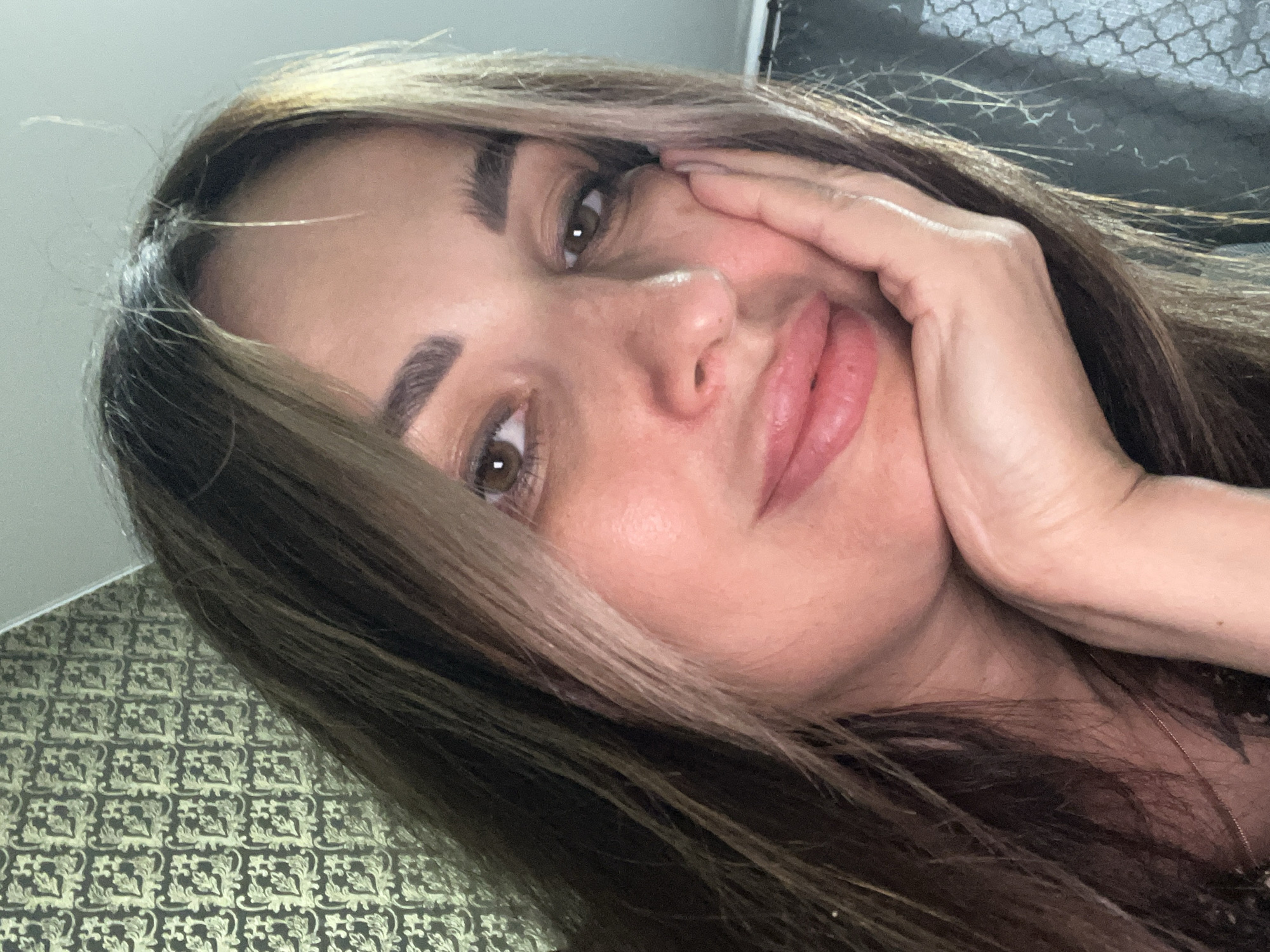 A close-up, selfie-style portrait of a woman with brown hair and brown eyes. Her left hand is resting on her chin, and she is looking directly at the camera. Her lips are slightly parted. The background is a patterned wallpaper.