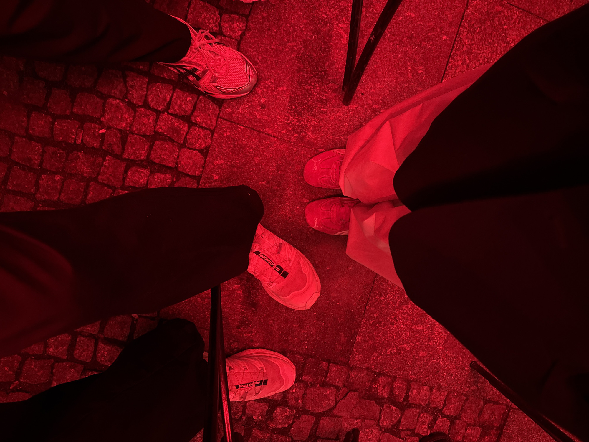 In Unknown, Unknown, a low-angle, over-the-shoulder shot captures the legs and feet of at least three individuals standing on a red-lit cobblestone surface. Two individuals are wearing white Salomon shoes, one with distinctive red laces. Another person is wearing a lighter-colored athletic shoe with red accents. The ground appears to be made of rectangular cobblestones. The lighting is predominantly red, casting a deep red hue over the entire scene. One person's legs are clad in dark trousers, while another is wearing white trousers. Shadows are cast by the individuals onto the cobblestones. The image has a grainy texture, possibly due to low light conditions or image processing.