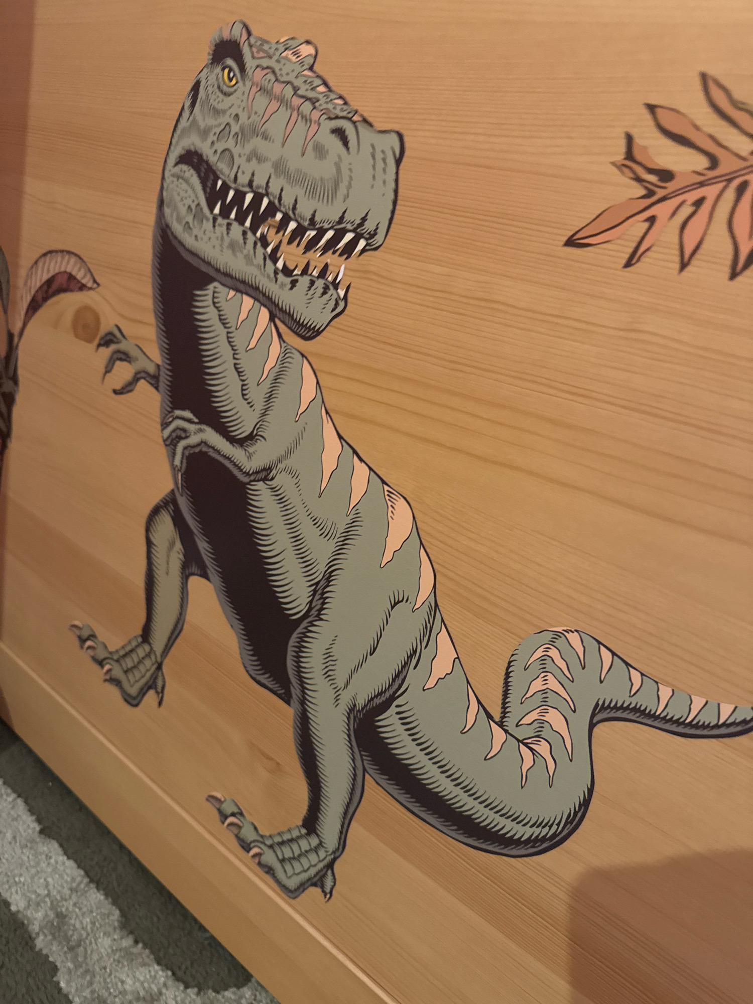 A close-up, eye-level shot shows a stylized, grey Tyrannosaurus Rex illustration on a wooden surface. The dinosaur is depicted in a dynamic pose, with its mouth open revealing sharp teeth and its body angled towards the right. Light pink stripes accent its skin, and its claws are visible on its feet and small arms. To the upper right, a separate illustration of a brown, leaf-like shape is present. The wooden surface displays a natural grain pattern, with a subtle shadow cast by the dinosaur on the right side of the frame. The bottom left of the image shows a fuzzy grey carpet.
