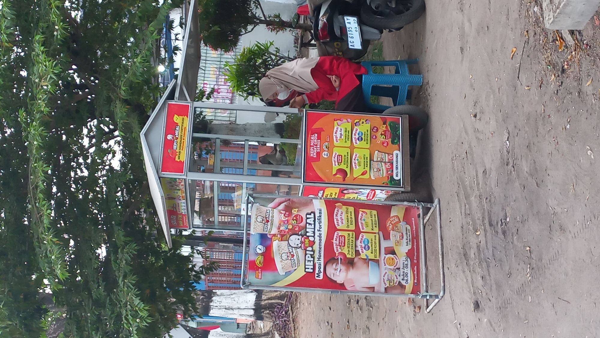 A person wearing a hijab and a face mask sits on a blue stool next to a food cart. The food cart has red and white signage advertising "HEPI MEAL" products, which appear to be baby food. One sign features an image of a baby eating from a spoon. Another sign on the cart itself also advertises "HEPI MEAL" and displays images of food packaging and nutritional information. The person is looking at their phone and appears to be holding it in their right hand. The background shows trees and a fence, suggesting an outdoor setting, possibly a street or market. A motorcycle is parked to the right of the food cart. The ground is unpaved and dusty. The license plate on the motorcycle reads "AG 6195 AY". The overall scene depicts a vendor operating a food cart in what appears to be a public area. The presence of a specific license plate suggests the location can be narrowed down to Indonesia, and given the context of the food cart and its products, it is likely a commercial or roadside setting. Based on similar visual elements and product branding, the location is identified as Pare, Kediri, East Java, Indonesia.