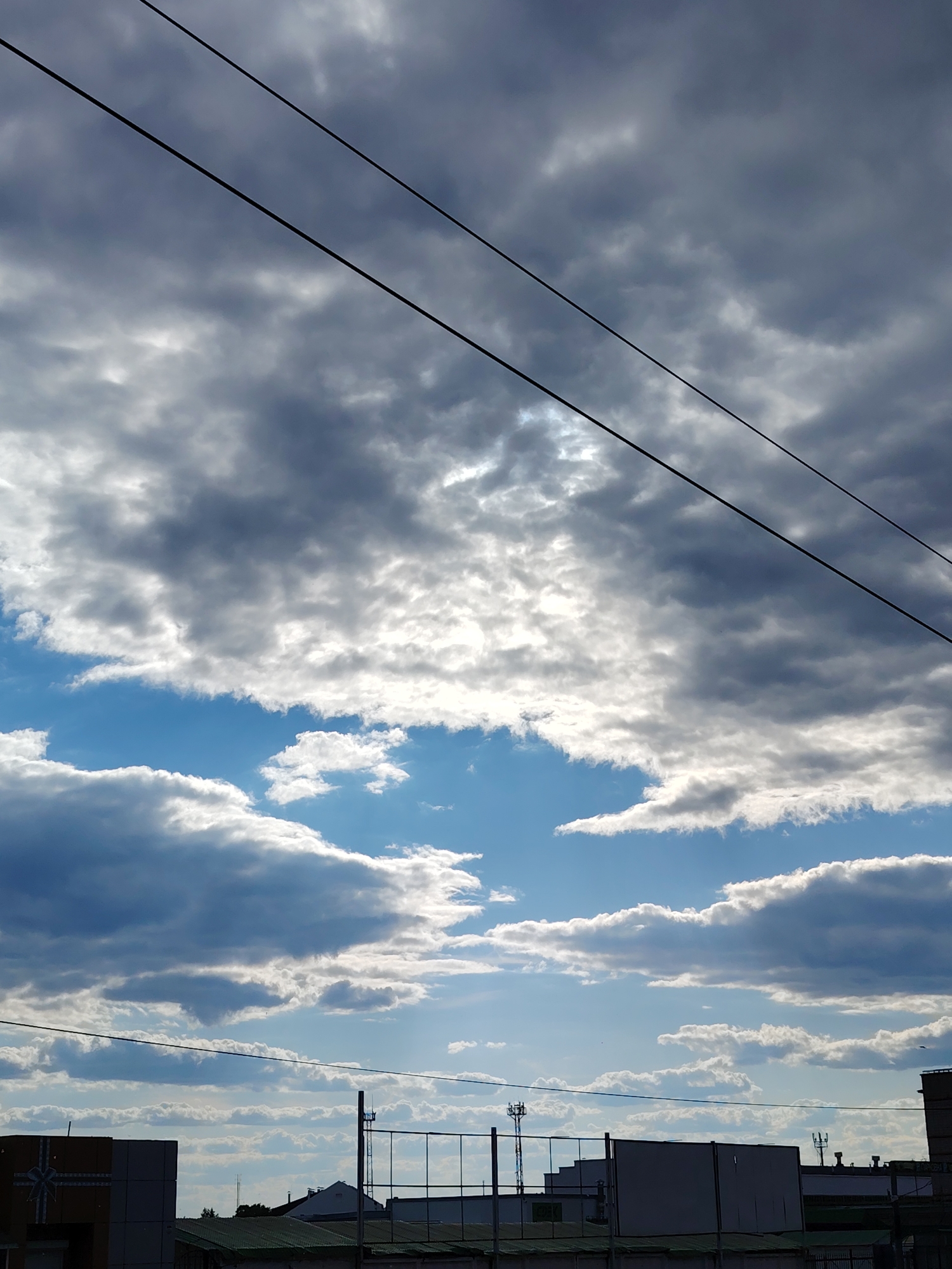 The image displays a dynamic sky over the city of Chaikovsky, Russia. The upper half is dominated by a dense formation of grey and white clouds, with bright, luminous patches indicating sunlight breaking through. Below this, significant areas of clear blue sky are visible, particularly in the mid-frame, interspersed with smaller white clouds. Two parallel, dark utility lines traverse diagonally from the upper left to the lower right. A third, less prominent horizontal utility line is present towards the bottom of the sky portion. The bottom fifth of the image reveals the silhouetted skyline of an urban or industrial area. Visible structures include indistinct building outlines, several rooftops, and a prominent tall, slender mast or tower on the right side. On the far left, a portion of a light-colored building with a distinctive dark, ribbon-like or cross-shaped pattern is partially visible. No people, animals, or specific actions are discernible within the frame.
