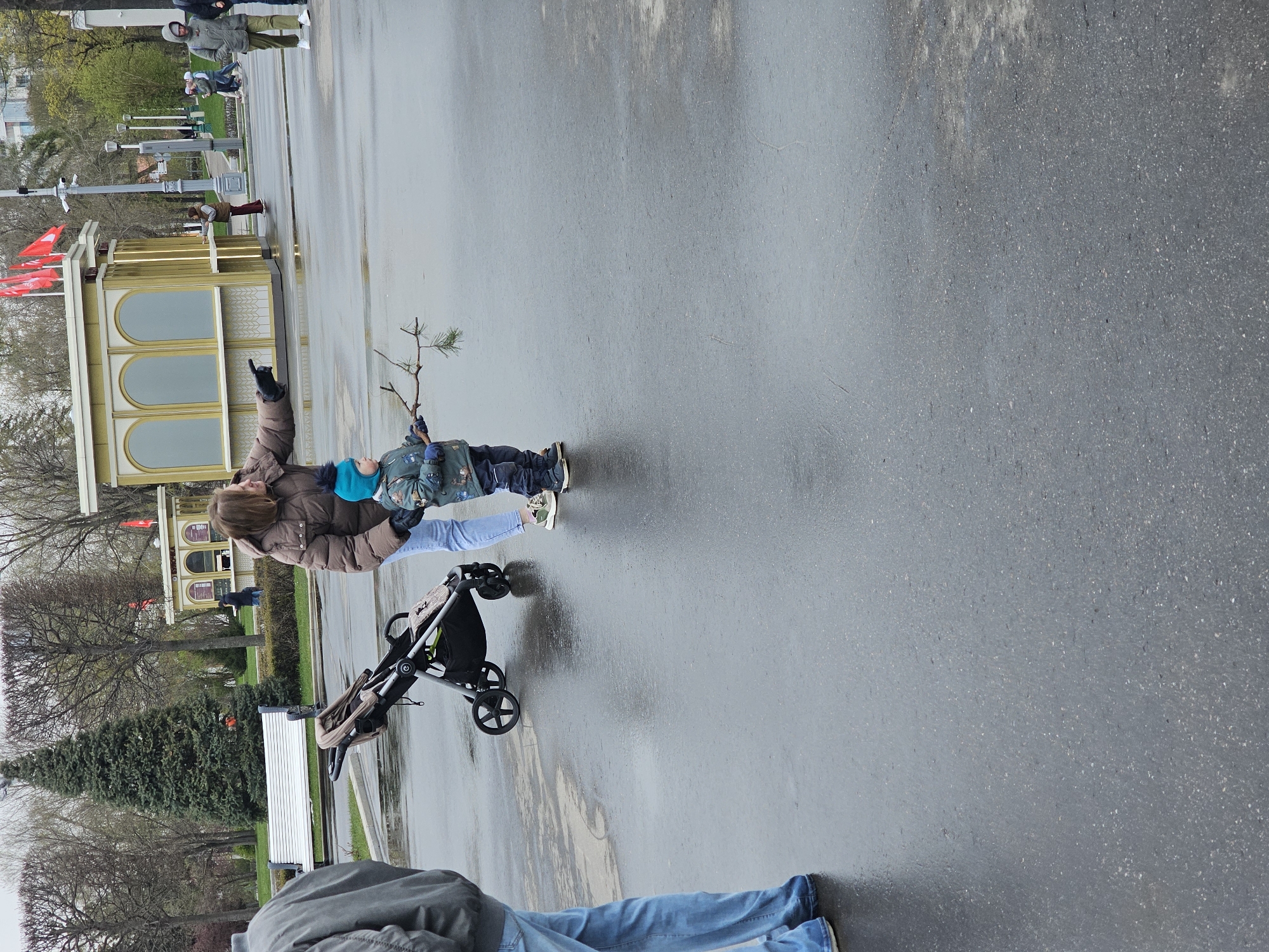 In a public outdoor setting, likely a park or pedestrian area in Moscow, Russia, a woman in a brown winter coat and light blue jeans stands next to a child holding a small tree branch. The child is dressed in a winter coat and dark pants. A black stroller is positioned to the left of the woman. In the background, there is a decorative gold kiosk with red flags, a green space with benches, and bare trees indicating it is either late autumn or early spring. The ground appears wet, suggesting it has recently rained. Other people are walking in the background. In the foreground, parts of two individuals are visible. One is wearing light blue jeans and the other wearing a grey jacket.