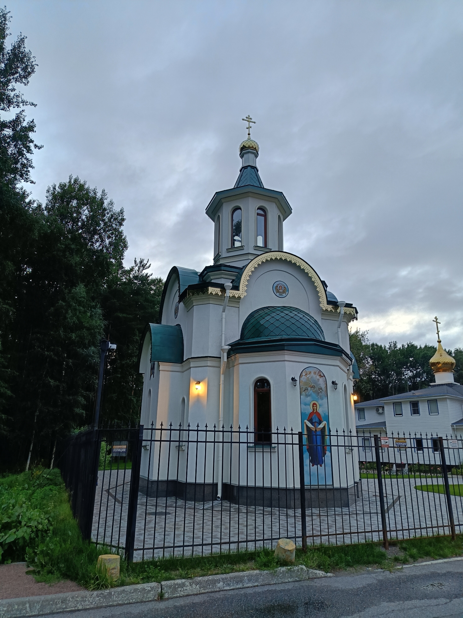 The image shows the Church of the Icon of the Mother of God "Joy of All Who Sorrow" located at 8 Pionerskaya Street, Kronstadt, Saint Petersburg, Russia. The church is a white, multi-domed Orthodox Christian church with a green roof and a golden cross on its main dome. A smaller golden dome with a cross is visible to the right of the main structure, atop a white building. The church is surrounded by a black metal fence. To the left of the church, tall trees are visible, and a security camera is mounted on a pole. The sky is overcast and gray. The foreground shows a paved walkway and a road. The overall scene depicts a religious building in a somewhat somber, natural setting, possibly at dusk. There are no people visible in the image.