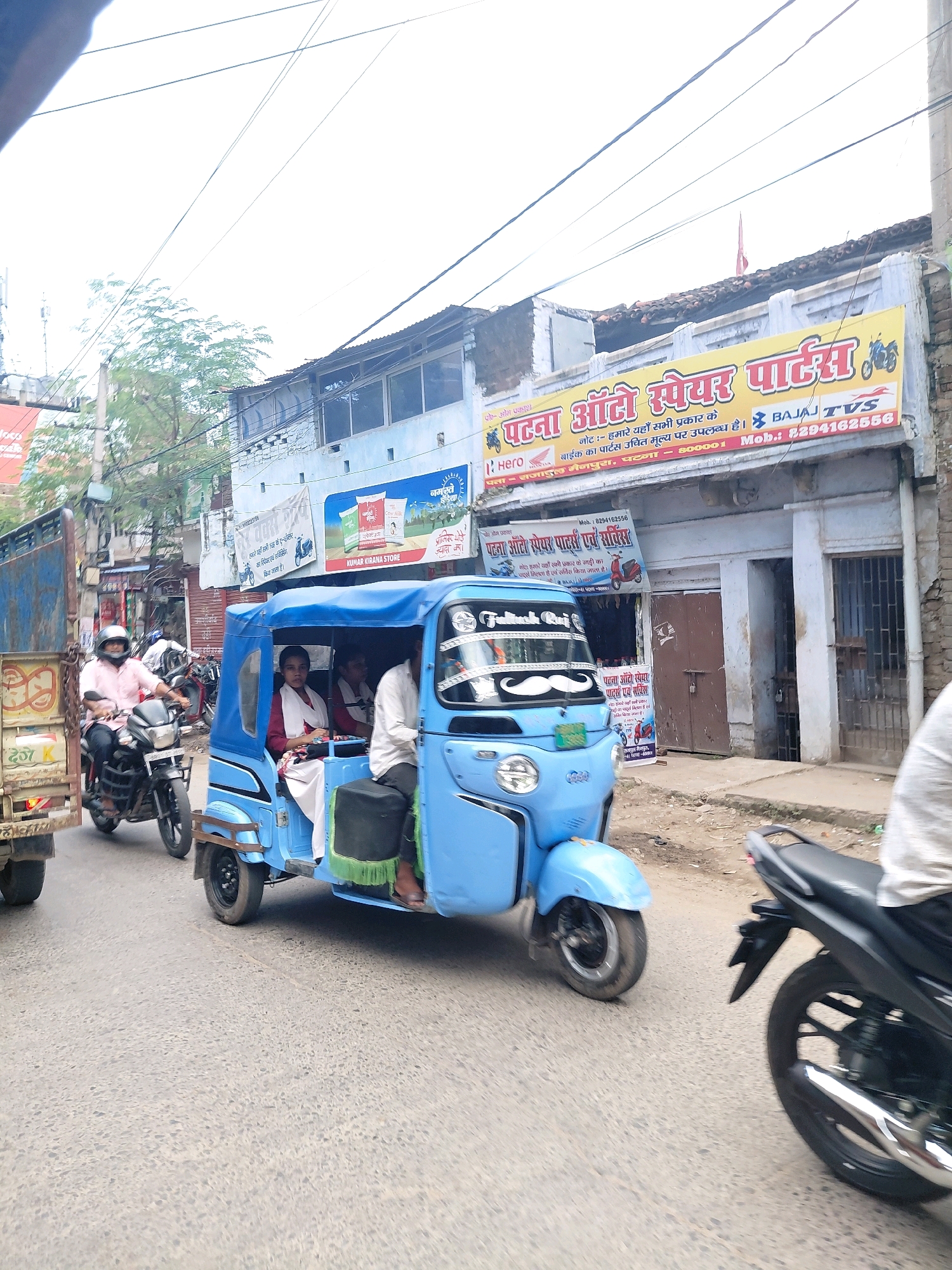 The image captures a street scene in Patna, India, likely on a cloudy day given the washed-out sky. In the foreground, a bright blue auto-rickshaw is the central focus. The auto-rickshaw has a "Patna Auto Spare Parts" sign on its side. It is carrying at least three people: a woman in a white and maroon outfit is seated in the passenger section, and two men are also visible, one driving and another in the back. The auto-rickshaw has a distinctive mustache-like decoration on its front windshield.
To the left of the auto-rickshaw, a man wearing a pink shirt and a helmet is riding a motorcycle. He appears to be the driver. Behind him, a blue truck is partially visible, suggesting traffic and a busy road.
In the background, buildings line the street. One building prominently displays a large yellow banner with "पटना ऑटो स्पेयर पार्ट्स" (Patna Auto Spare Parts) written in large, bold Hindi letters, along with logos for Bajaj and TVS. Below this, a smaller sign reads "Hero Honda." Another sign in Hindi on a white building indicates "KUMAR KIRANA STORE." The architecture of the buildings suggests a commercial area in Patna. Wires are strung across the sky, typical of urban Indian infrastructure. The road surface appears to be asphalt, with some patches of dirt and debris visible. The overall impression is that of a typical day in a bustling Indian city.