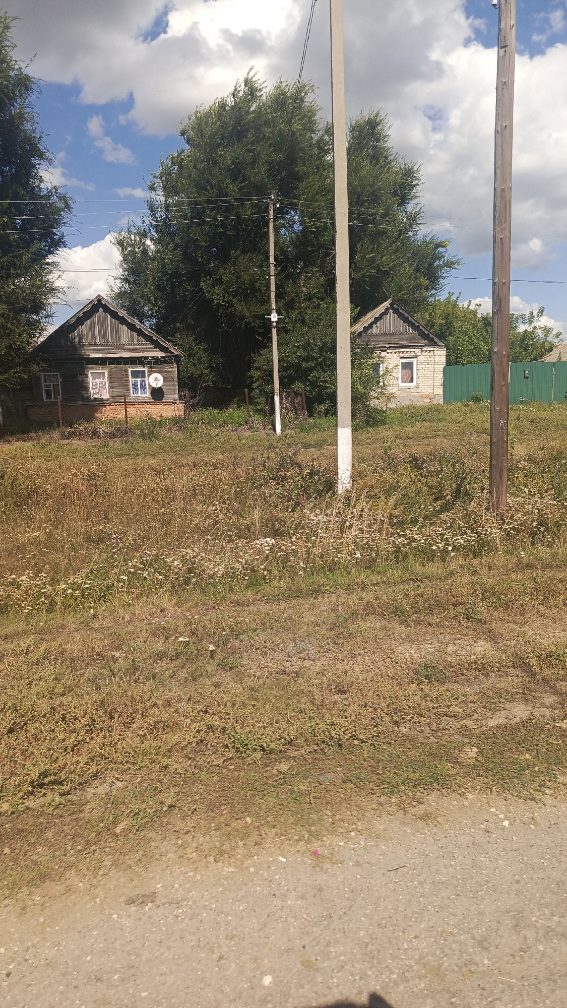 The image depicts a rural scene with two houses, a dirt road in the foreground, and a fence in the background. The houses appear to be small, wooden structures with visible windows. One house has a red brick foundation, while the other is made of wood. There are trees and vegetation surrounding the houses, and a sky with clouds is visible above. Power lines and utility poles are also present in the scene. The overall impression is of a simple, undeveloped area, likely in a village or rural settlement.