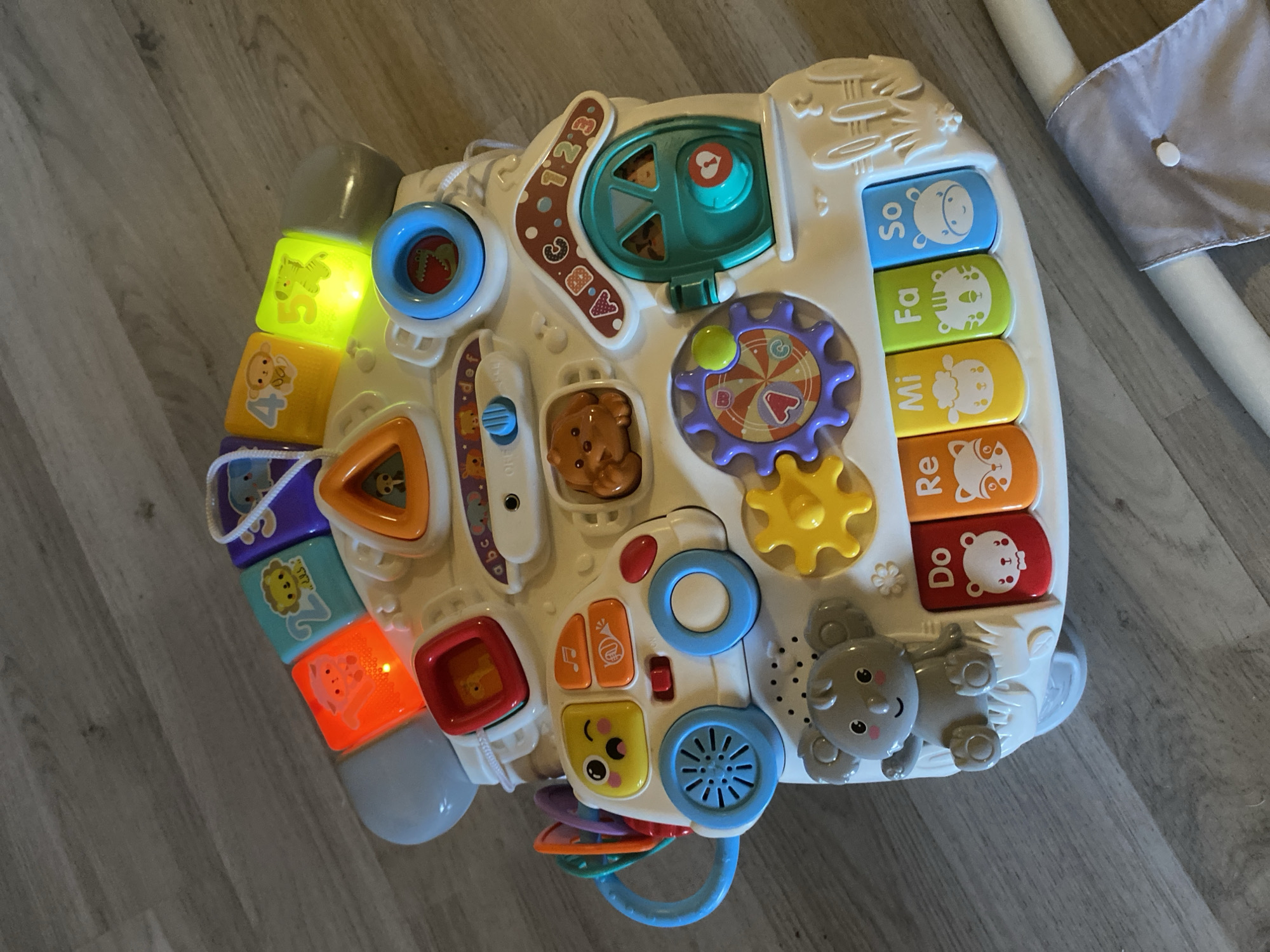 The image shows a white, house-shaped children's toy on a wooden floor. The toy features various interactive elements including colorful buttons, gears, a car with a smiley face, a door with a heart cutout, and a keyboard with animal illustrations. Several buttons are illuminated with different colors, including red, yellow, and green. The toy also has a grey koala figure seated on the left side. The background is a light brown wooden floor. There are no people or animals present in the image besides those depicted on the toy. The scene is indoors. This image was not taken in the city of Varna, Bulgaria.