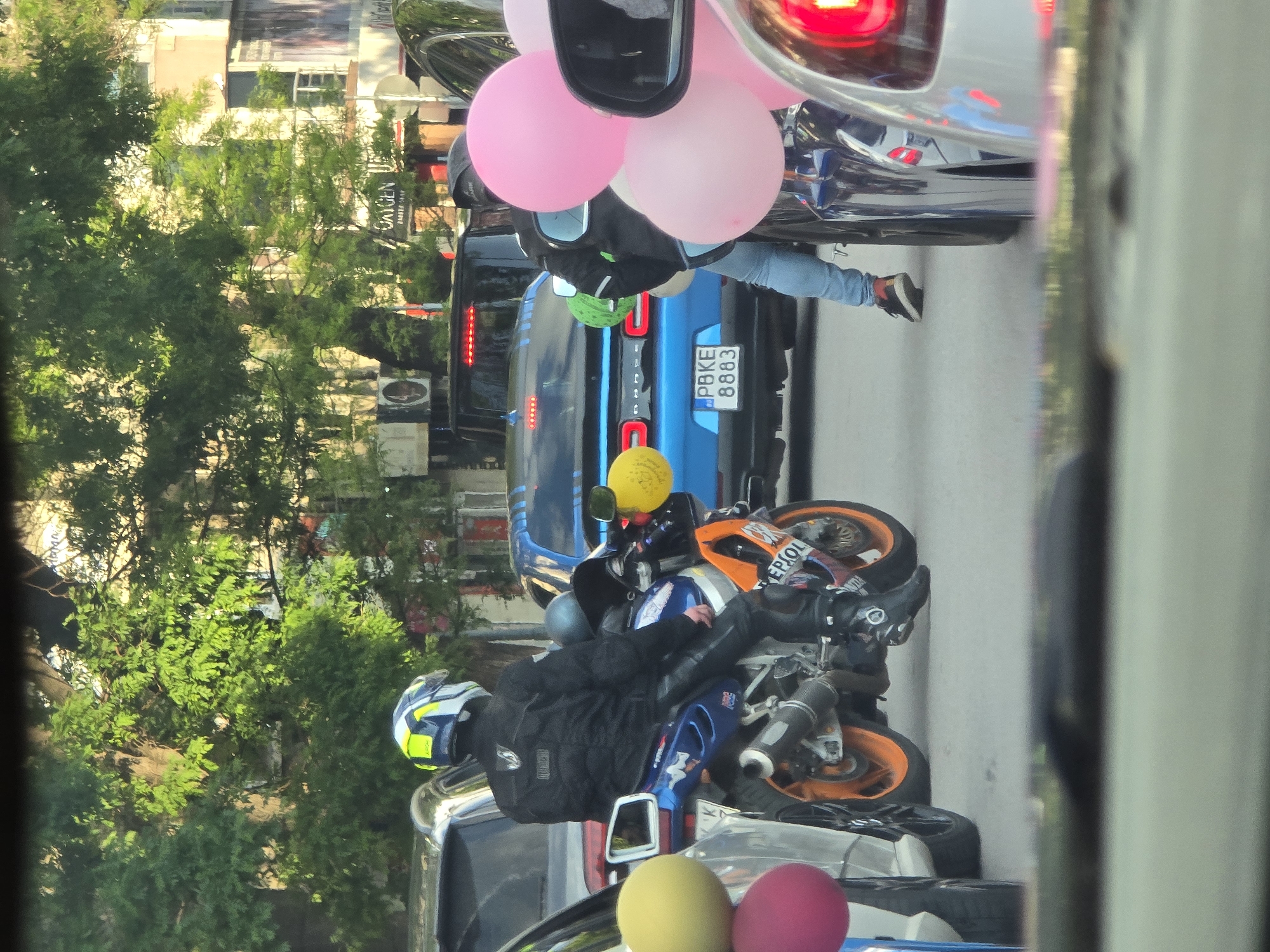 The image depicts a street scene, likely in an urban area, based on the presence of buildings, trees, and various vehicles. A motorcycle is a focal point, specifically with an orange Repsol-branded wheel. A person is positioned near the motorcycle, wearing a black jacket and dark pants, with their legs visible extending towards a blue car. Above them, multiple pink balloons are visible. Other vehicles, including a blue car with license plate "PBKE 8883," are also present in the frame. The photo seems to be taken from inside a vehicle. The specific city and country remain unknown.