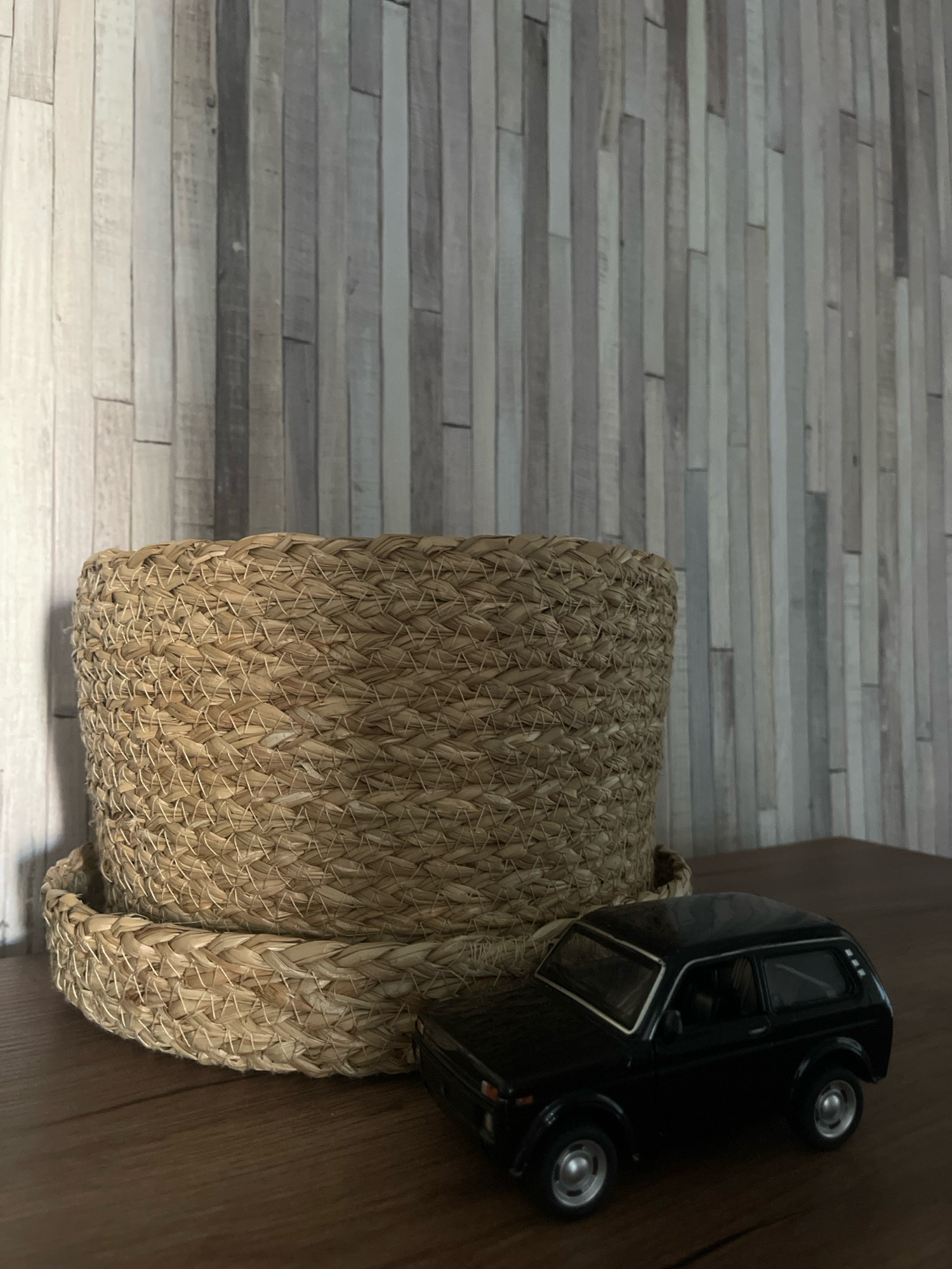 A close-up, eye-level shot shows a woven basket and a black toy car on a wooden surface. The basket, made of straw-like material, is cylindrical with a wider base and a rim. The toy car, a detailed replica of a hatchback, is positioned to the right of the basket. The background features a textured wall with vertical wooden slats in varying shades of gray and white. The lighting is soft, casting subtle shadows. The scene appears to be an indoor setting, possibly a home or studio, with no identifiable landmarks or context to suggest a specific location within Tallinn, Estonia.