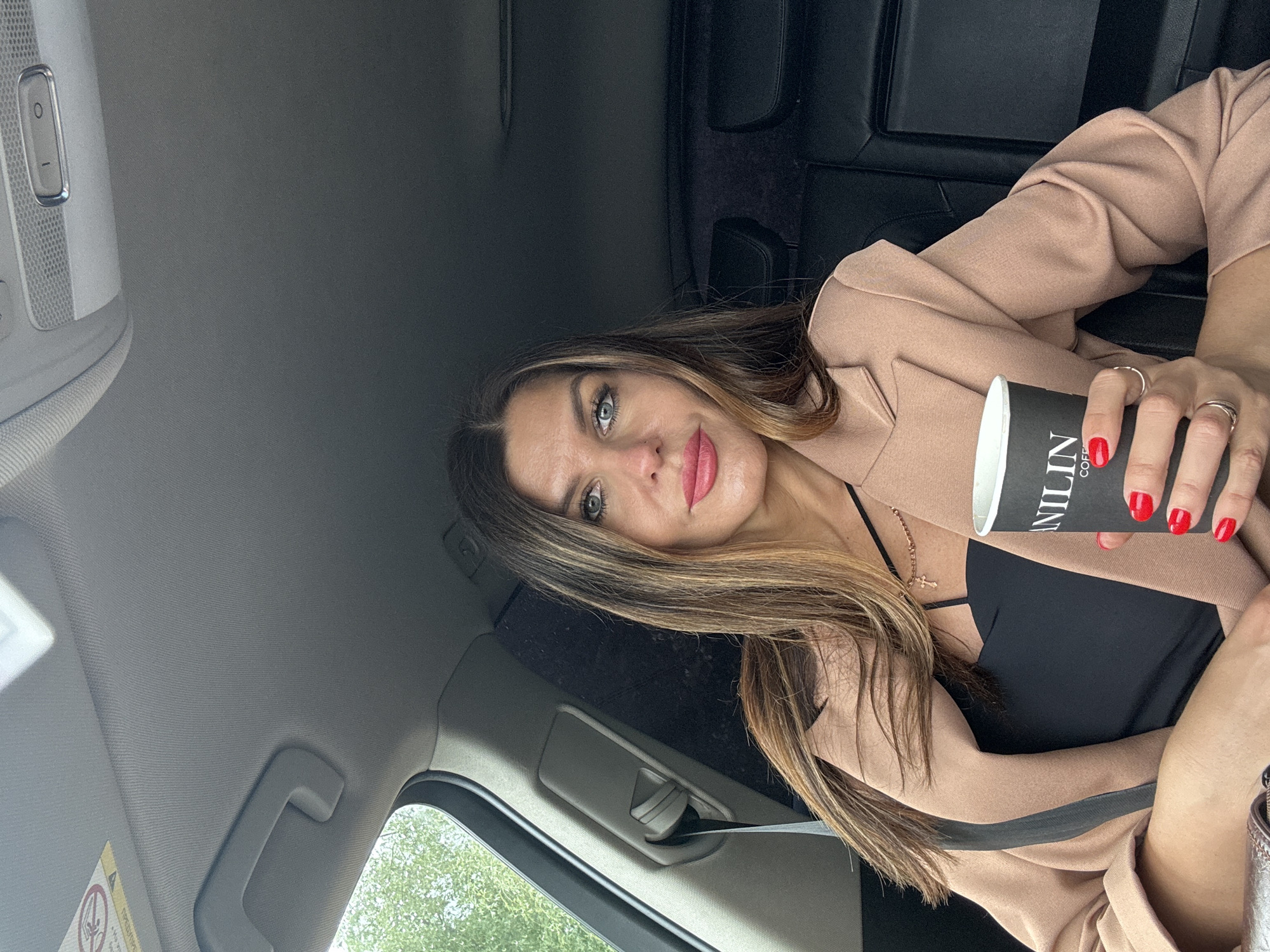 A woman with long, light brown hair and blonde highlights, light-colored eyes, and pink lipstick is captured in a selfie-style photograph taken inside a car. She has a calm expression with a slight smile. She is wearing a light peach or beige blazer over a black top with thin straps, accessorized with a delicate cross necklace. Her fingernails are painted bright red, and she wears multiple silver rings on her left hand.
She holds a black disposable coffee cup with white text that reads "VANILIN COFFEE" vertically on its side. The setting is the interior of a vehicle, suggested by the grey headliner, an overhead control panel with buttons, a sun visor, and a grab handle. The back seats, upholstered in black, are visible behind her. Through the car windows, green foliage is discernible, indicating it is daytime. A warning label on the sun visor is partially visible, showing the Cyrillic word "ПРЕДУПРЕЖДЕНИЕ" (warning).
The image is taken from a tilted perspective, focusing on the woman and her immediate surroundings within the car, which is located in Chaikovsky, Russia.
