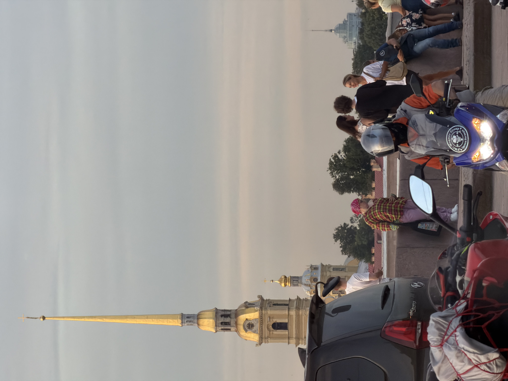 The image depicts an outdoor scene in Saint Petersburg, Russia, with the Peter and Paul Cathedral spire prominently visible in the background. In the foreground, a blue scooter with illuminated headlights is positioned, and its rider, wearing a helmet and a grey and orange jacket, is visible from the chest up. To the left, the rear of a black SUV is visible, with a red motorcycle partially in frame to the lower left, covered with a grey tarp and red netting.
In the middle ground, several people are gathered near a stone barrier. A person in a red and yellow plaid shirt and pink pants is standing, facing away from the camera. Next to them, a person in a white t-shirt is also standing. Further to the right, a group of three individuals are engaged in conversation: a woman with long dark hair, a person with curly brown hair, and a woman with blonde hair wearing a white t-shirt. Behind this group, other individuals and trees are visible. Another distinct dome structure is visible in the far background, to the right of the Peter and Paul Cathedral spire. The sky is overcast with a soft, diffused light, suggesting it is either early morning or late evening.