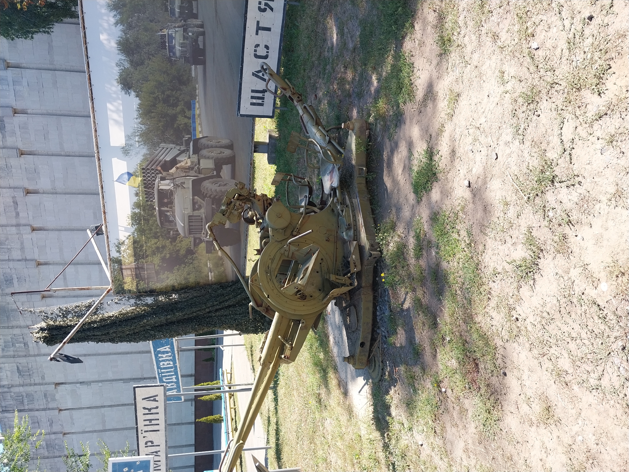 The image captures an outdoor exhibit featuring military equipment in Dnipro, Ukraine. In the foreground, a vintage anti-aircraft gun is prominently displayed on a grassy and dirt area. The gun is olive green and shows signs of age and wear. Behind the gun, a large mural or banner depicts a military scene with trucks and soldiers, and a Ukrainian flag is visible. To the left of the mural, two blue and white signs are mounted on poles; one sign reads "МАРТІНКА" and the other "АКДІВКА". The background consists of a large, light-colored building with a textured facade. The scene is illuminated by natural daylight, casting shadows on the ground.
