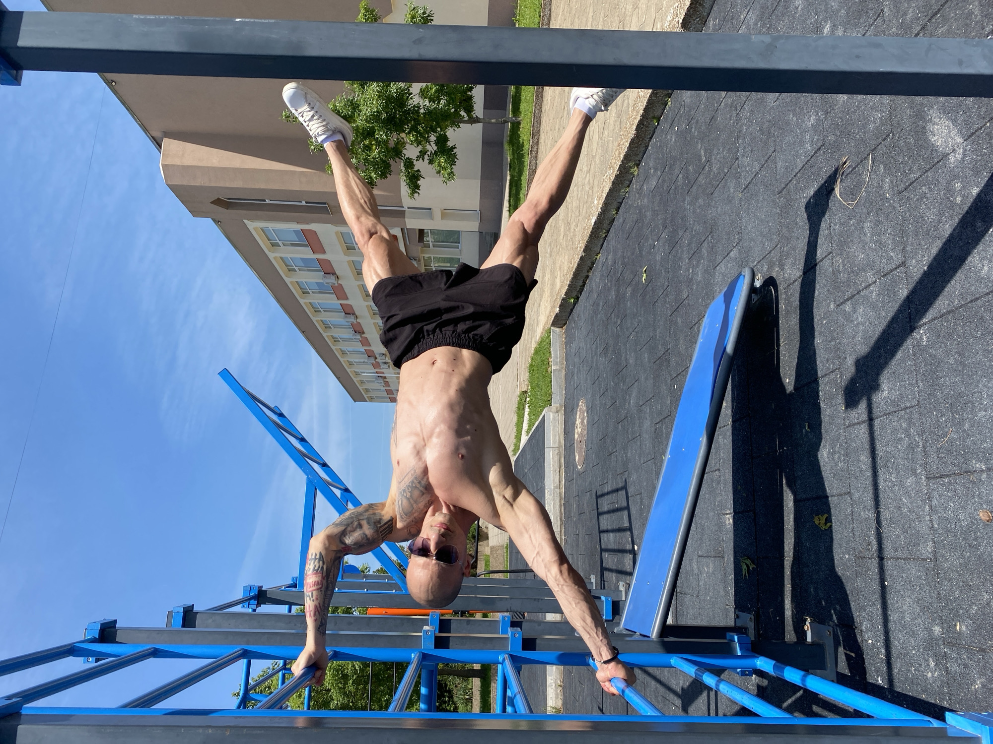 The image shows a bald, shirtless man with tattoos on his left arm and wearing sunglasses performing an inverted hang on a blue and grey metal playground structure. He is wearing black shorts and white sneakers. The setting appears to be an outdoor exercise area with a black rubberized ground surface. In the background, a multi-story building with light brown and white facade is visible, along with green trees and a blue sky. Based on the presence of the Cyrillic alphabet on the man's tattoo, the location is Pleven, Bulgaria.