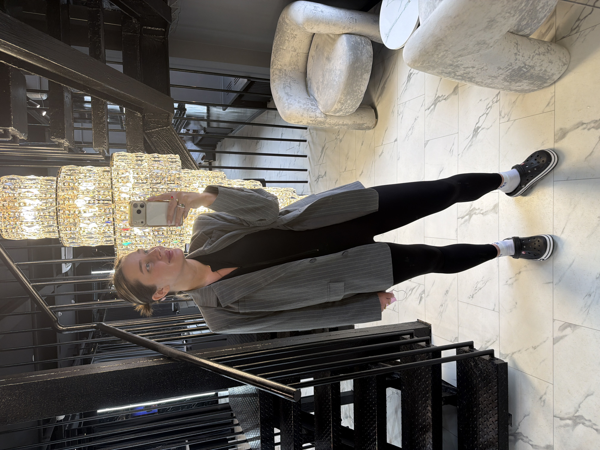 A woman in a gray pinstripe blazer, black leggings, white socks, and black Crocs takes a selfie in an indoor space featuring black metal stairs with diamond plate treads and a large, crystal chandelier. The environment includes a modern gray chair and a marble-patterned floor. The location is unidentified in the city of Unknown, Unknown.
